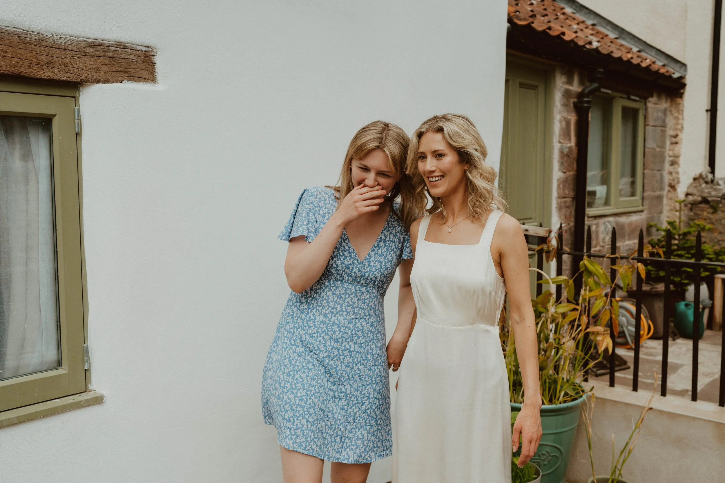 Two women standing outdoors, one wearing a blue dress with a floral pattern, laughing and covering her mouth, and the other wearing a white dress, smiling. They are near a white wall and a house with stone and brick elements, a window, and potted pla