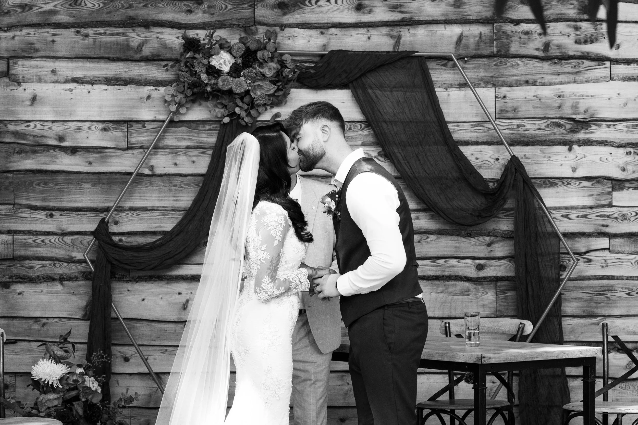 Black and white photo of a bride and groom kissing during their wedding ceremony in front of a wooden wall with decorative black fabric and flowers overhead.