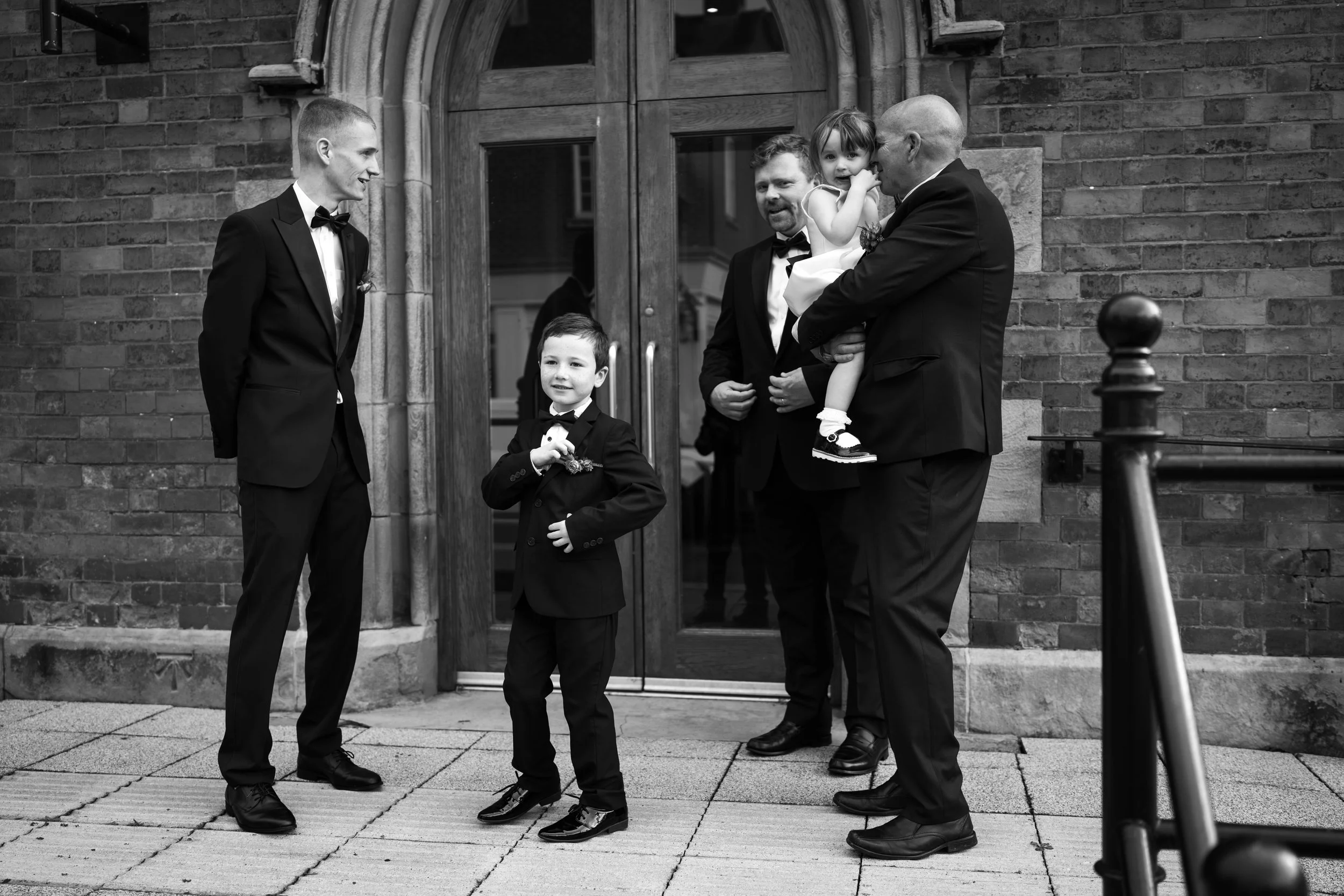 A group of five people dressed in formal attire standing outside a brick building, including two young boys and three men, one of whom is holding a young girl. They appear to be at a wedding or special event.