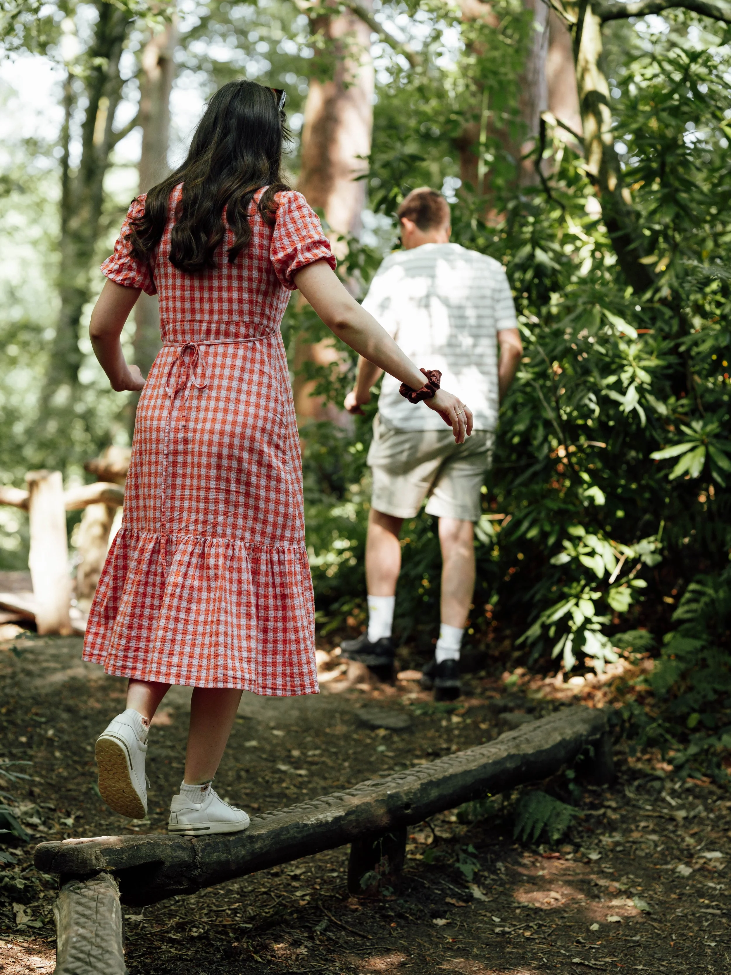 A woman in a red checkered dress and white sneakers balances on a narrow log on a forest trail while a man in a striped shirt and shorts walks ahead among green trees.