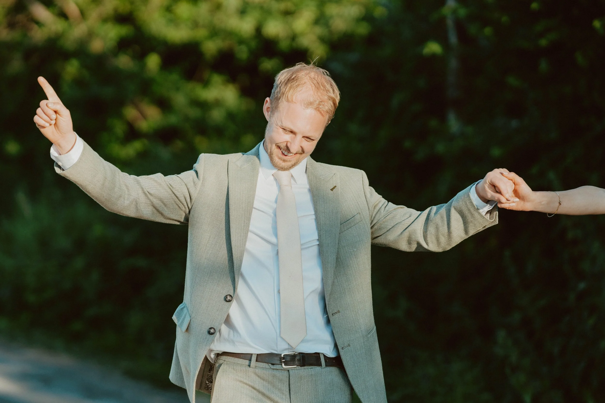A man in a beige suit and tie smiling and holding hands with someone off-camera outdoors with greenery in the background.
