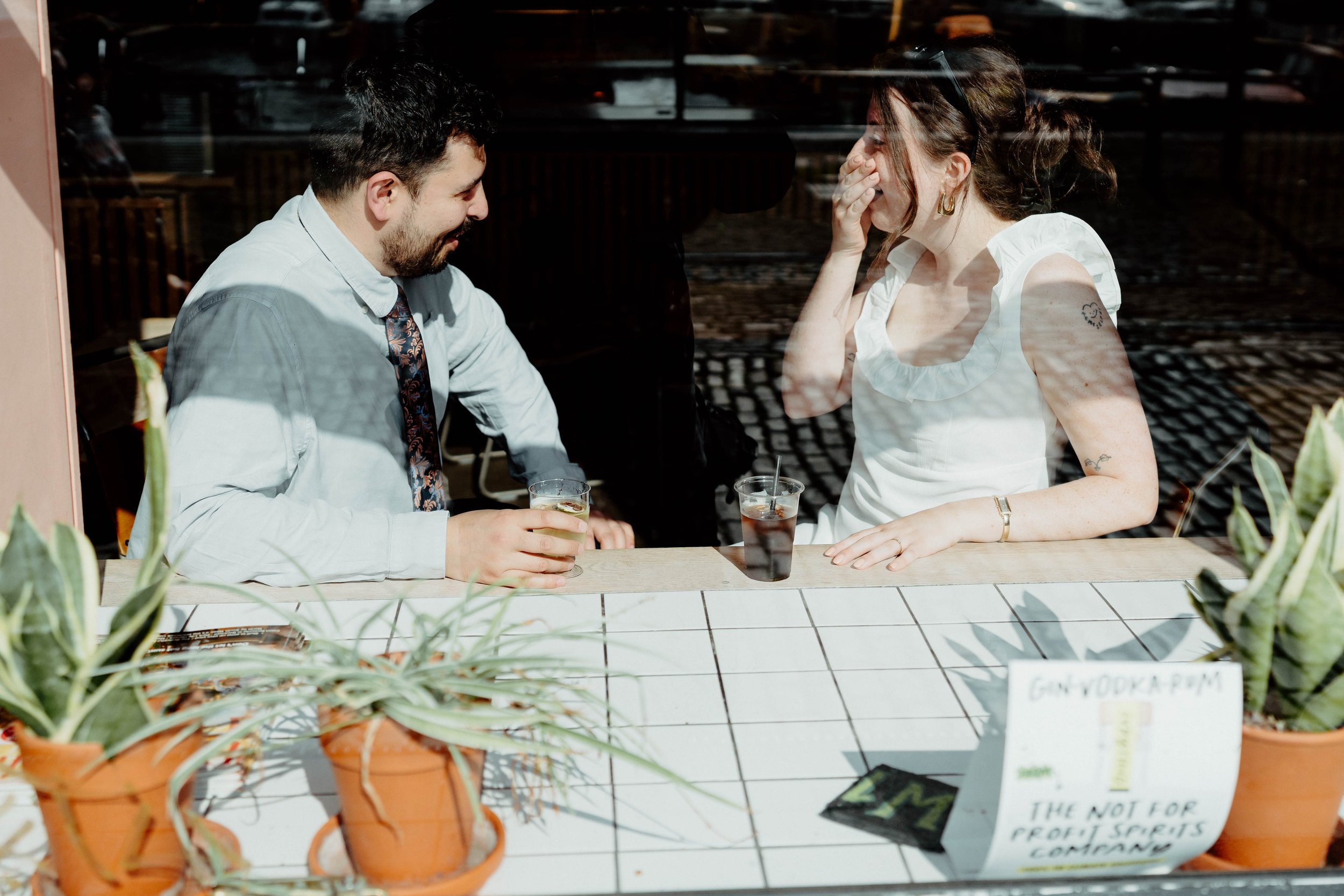 A man and woman are sitting at a restaurant window, sharing a laugh. The woman covers her mouth as she laughs, and both are holding drinks. Potted plants are on the table, with a sign that reads 'GANY DOKA PUM' and some printed text.