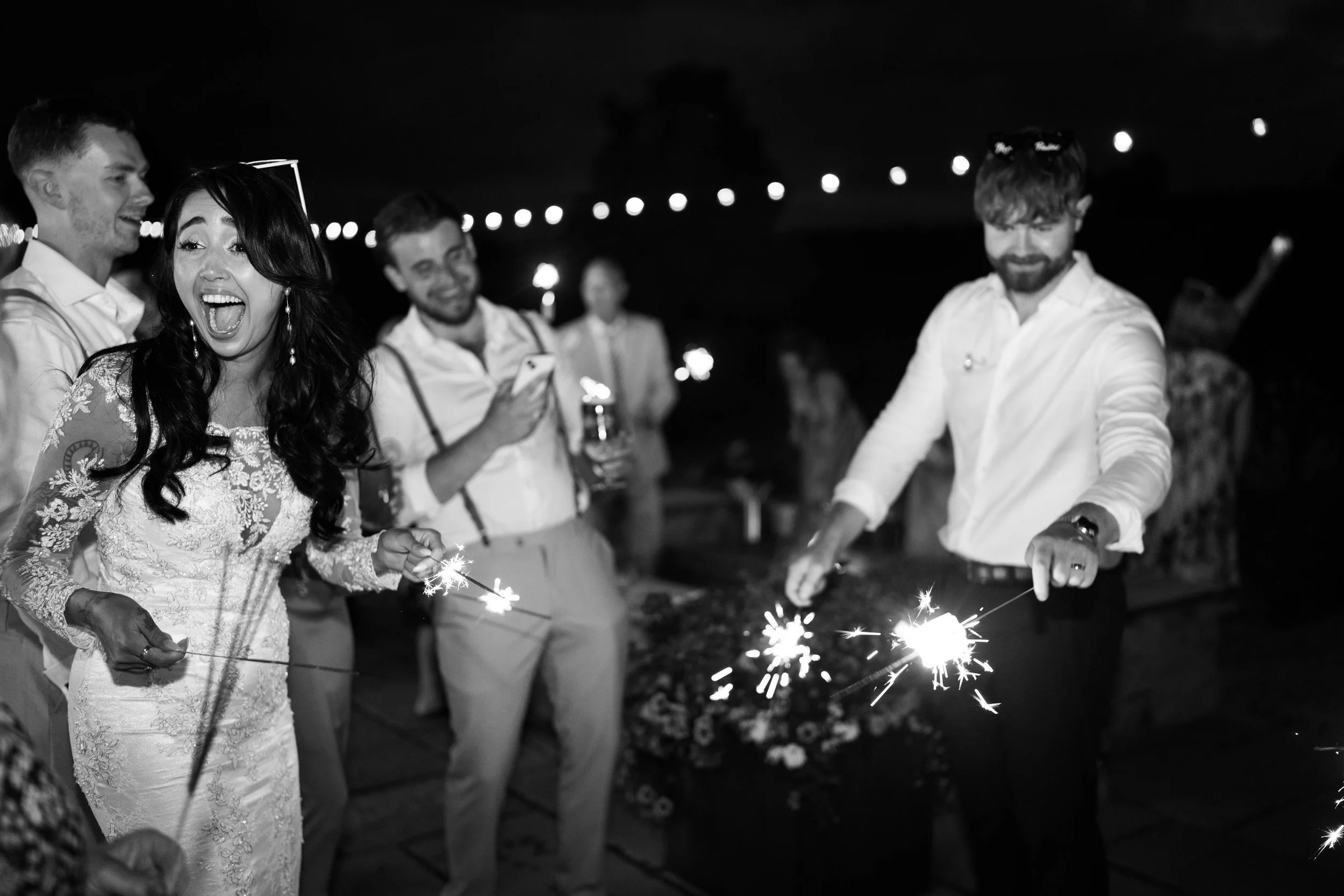 A group of people celebrating at a nighttime event, with some holding sparklers and others taking photos, smiling and enjoying the moment.