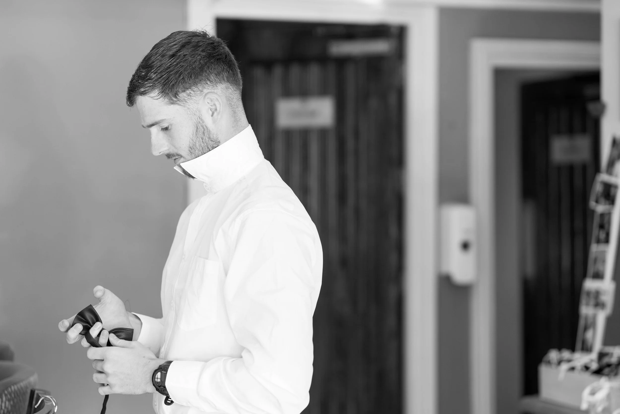 A young man in a white button-down shirt looking at a bow tie in his hands indoors.