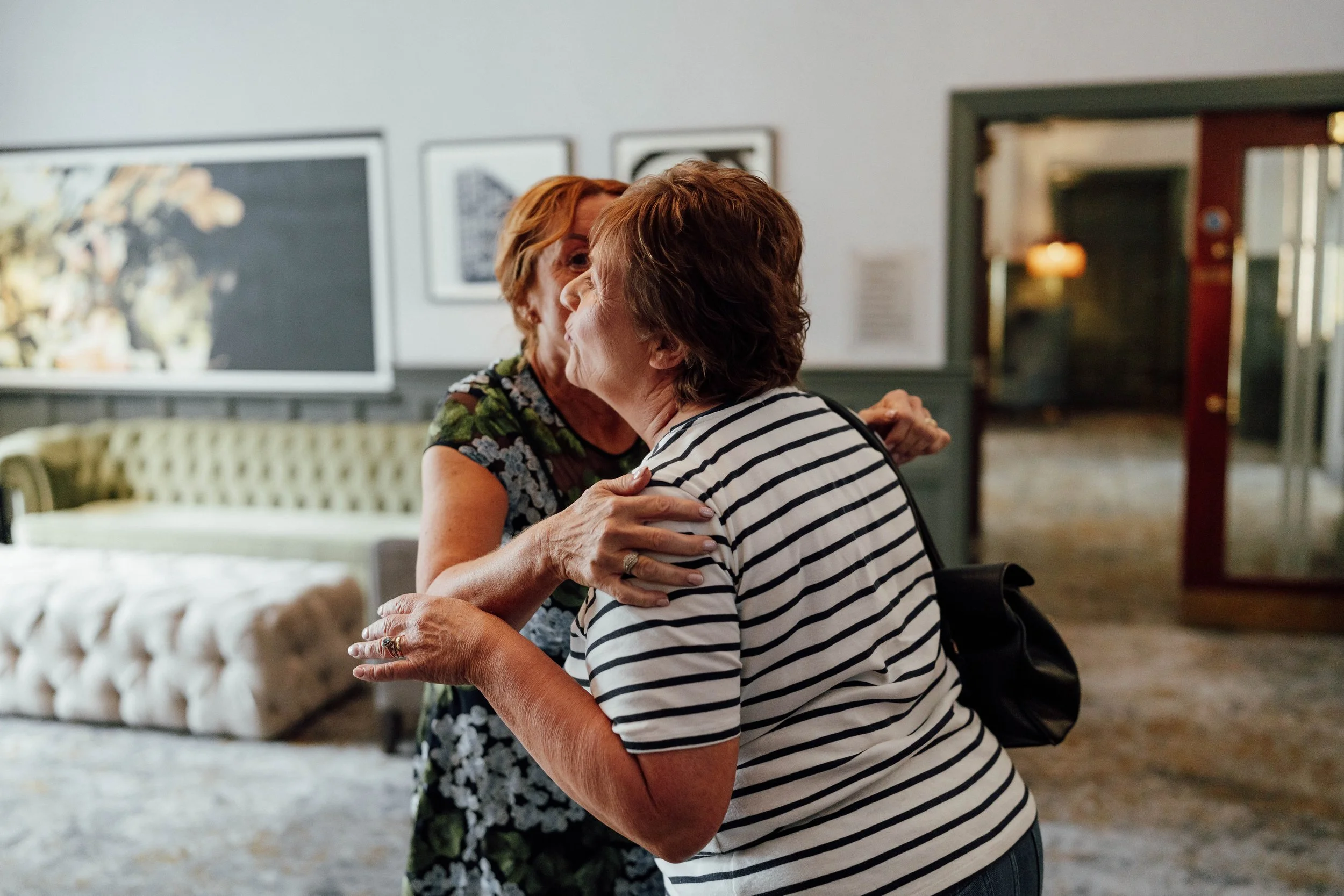 Two elderly women hugging and embracing warmly in a cozy, well-lit room, with artwork and a green velvet couch in the background.