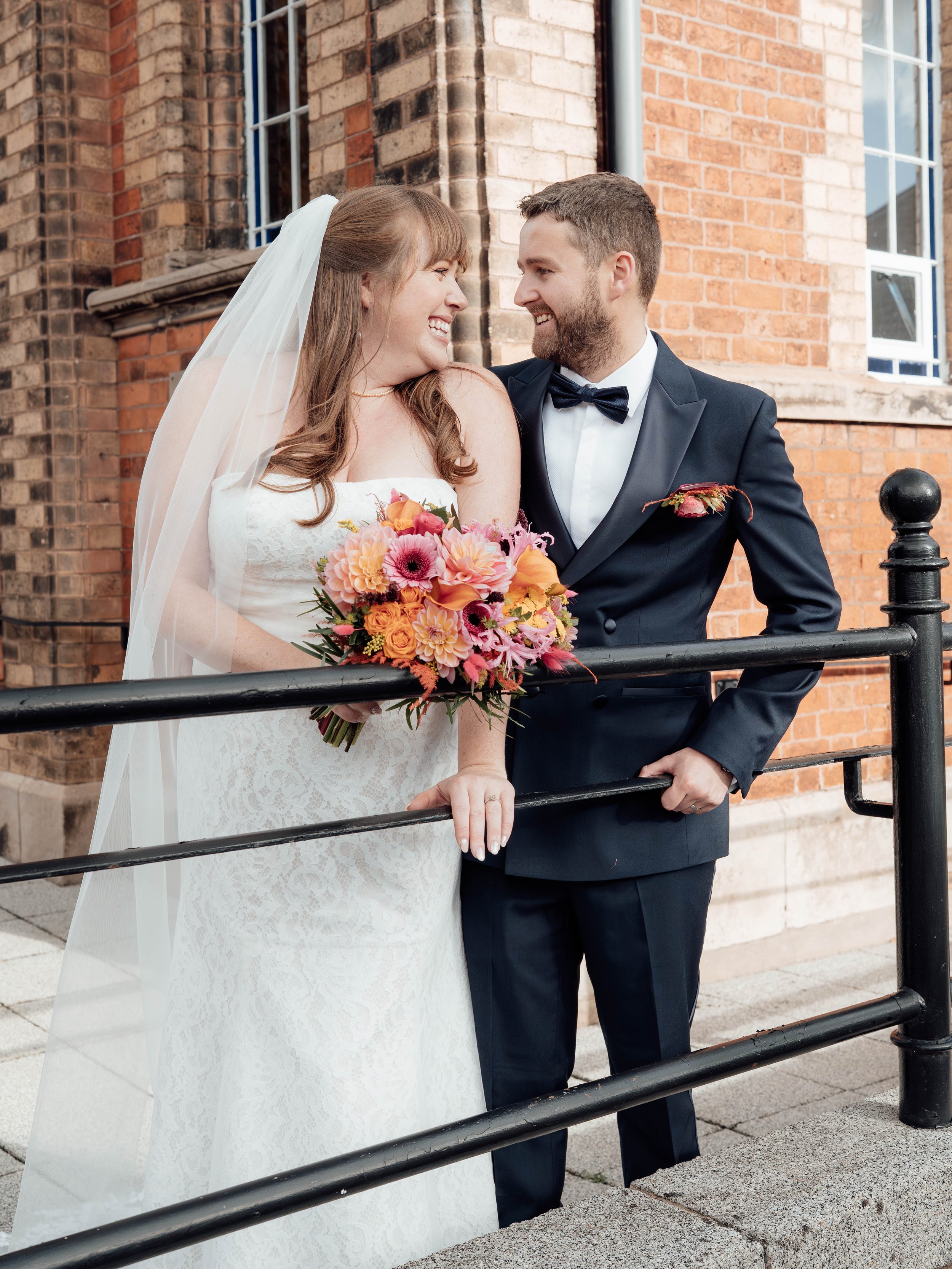 Bride and groom smiling at each other outside a brick building, with the bride holding a colorful bouquet of flowers.