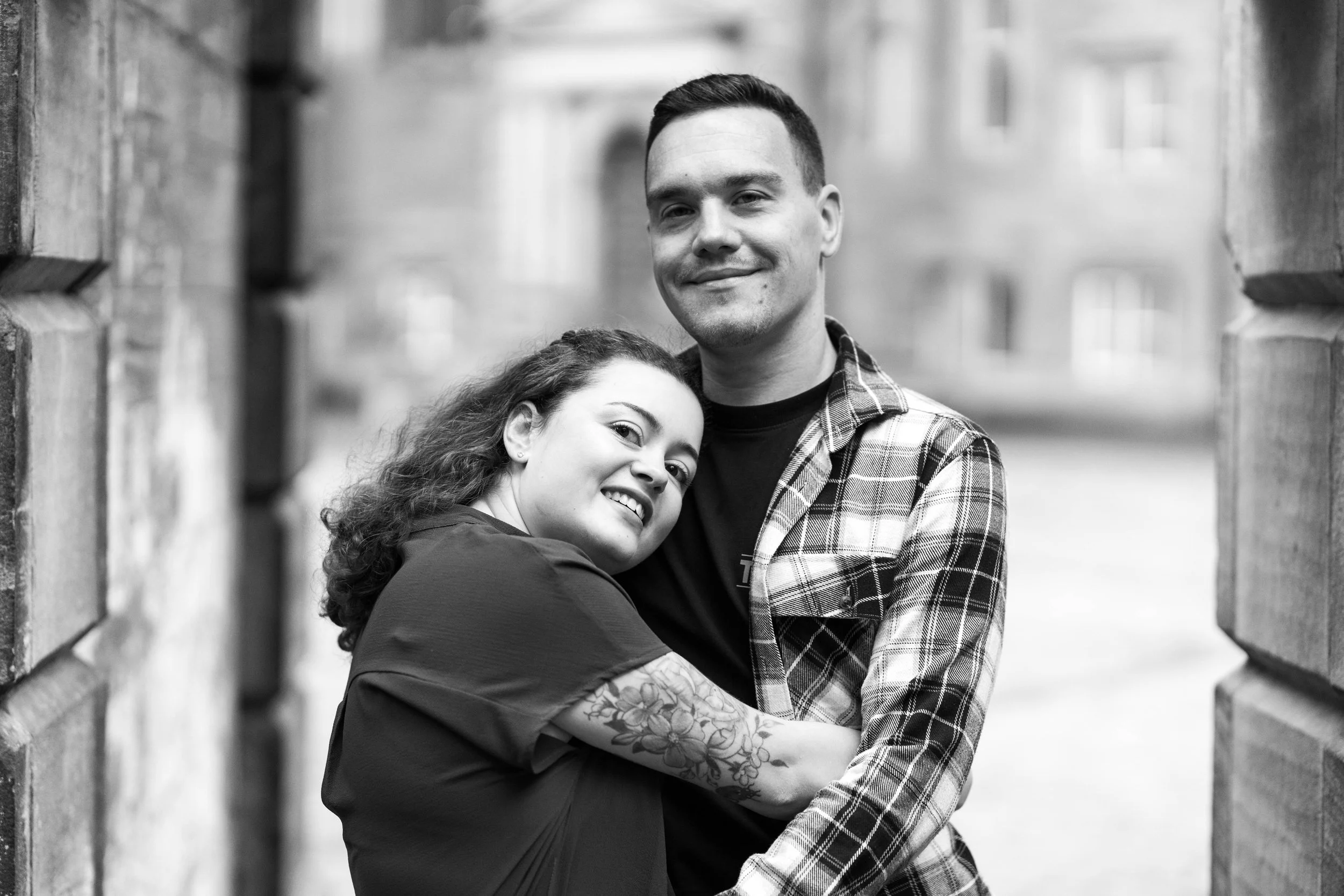 A black and white photo of a smiling couple embracing in an outdoor urban setting, with brick walls on either side.