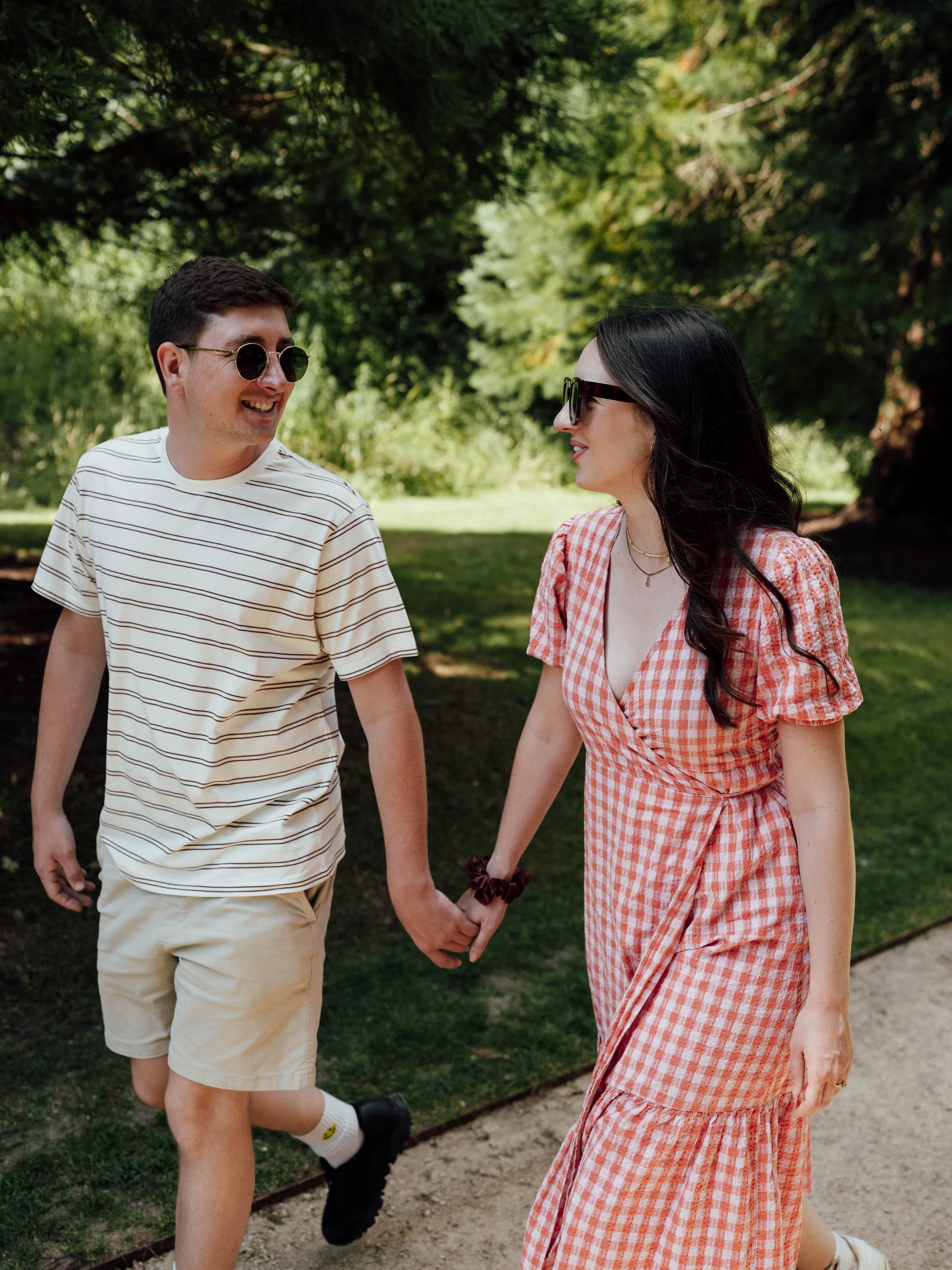 A couple holding hands and walking outdoors in a park on a sunny day. The man is wearing a striped T-shirt, beige shorts, and sunglasses. The woman is wearing a red and white checkered dress and sunglasses, with long dark hair.