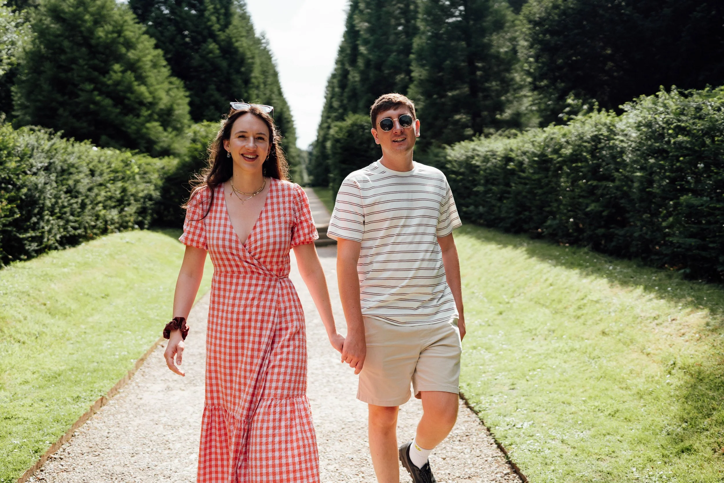 A young couple holding hands and walking along a gravel path in a lush, green park with tall trees on either side, smiling and enjoying a sunny day.