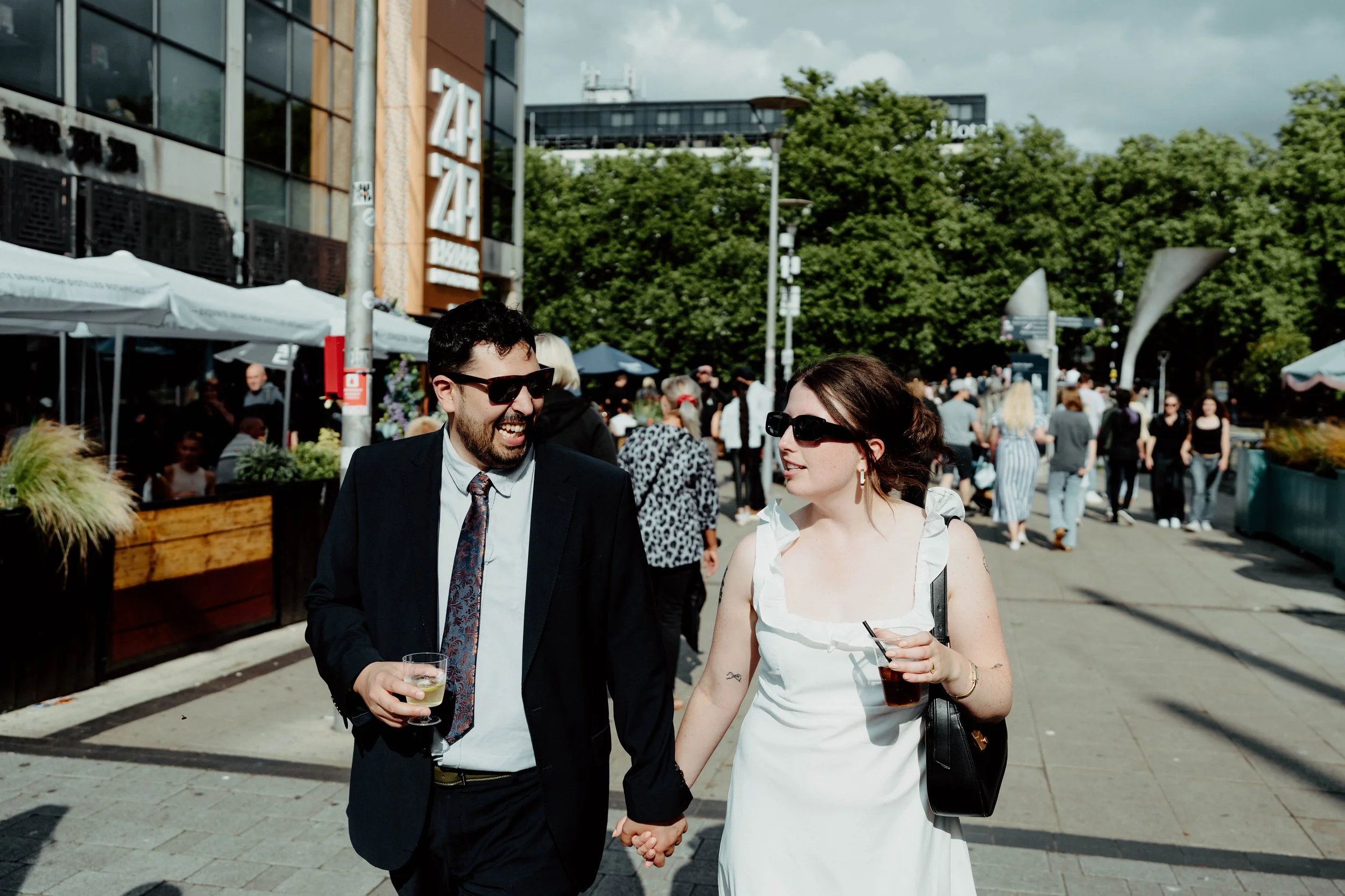 A man and woman in formal attire walking and holding hands outdoors, enjoying drinks on a sunny day. They are surrounded by other people, tents, and trees in an urban setting.