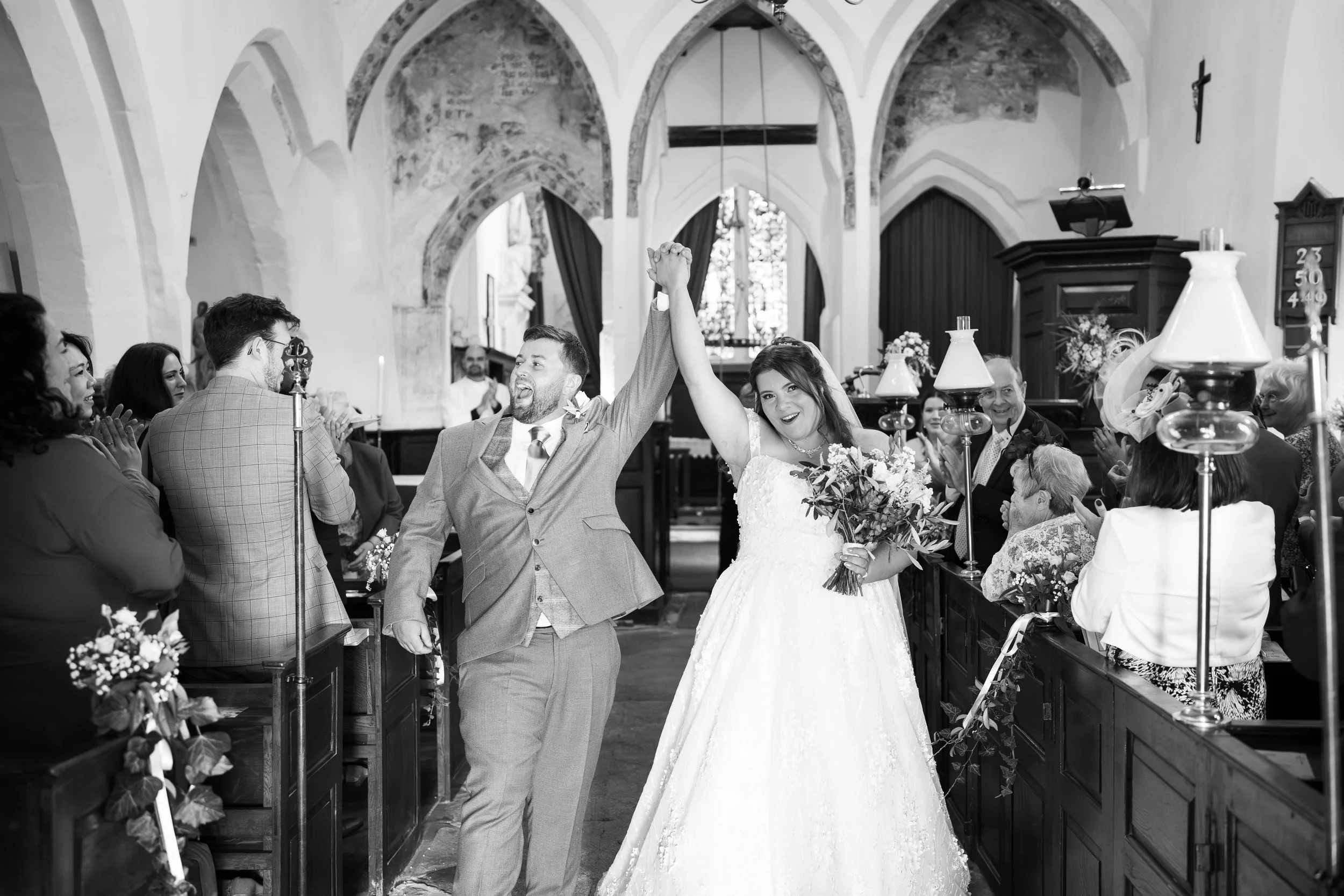 Black and white photo of a bride and groom celebrating wedding in a church, holding hands up high, surrounded by guests.