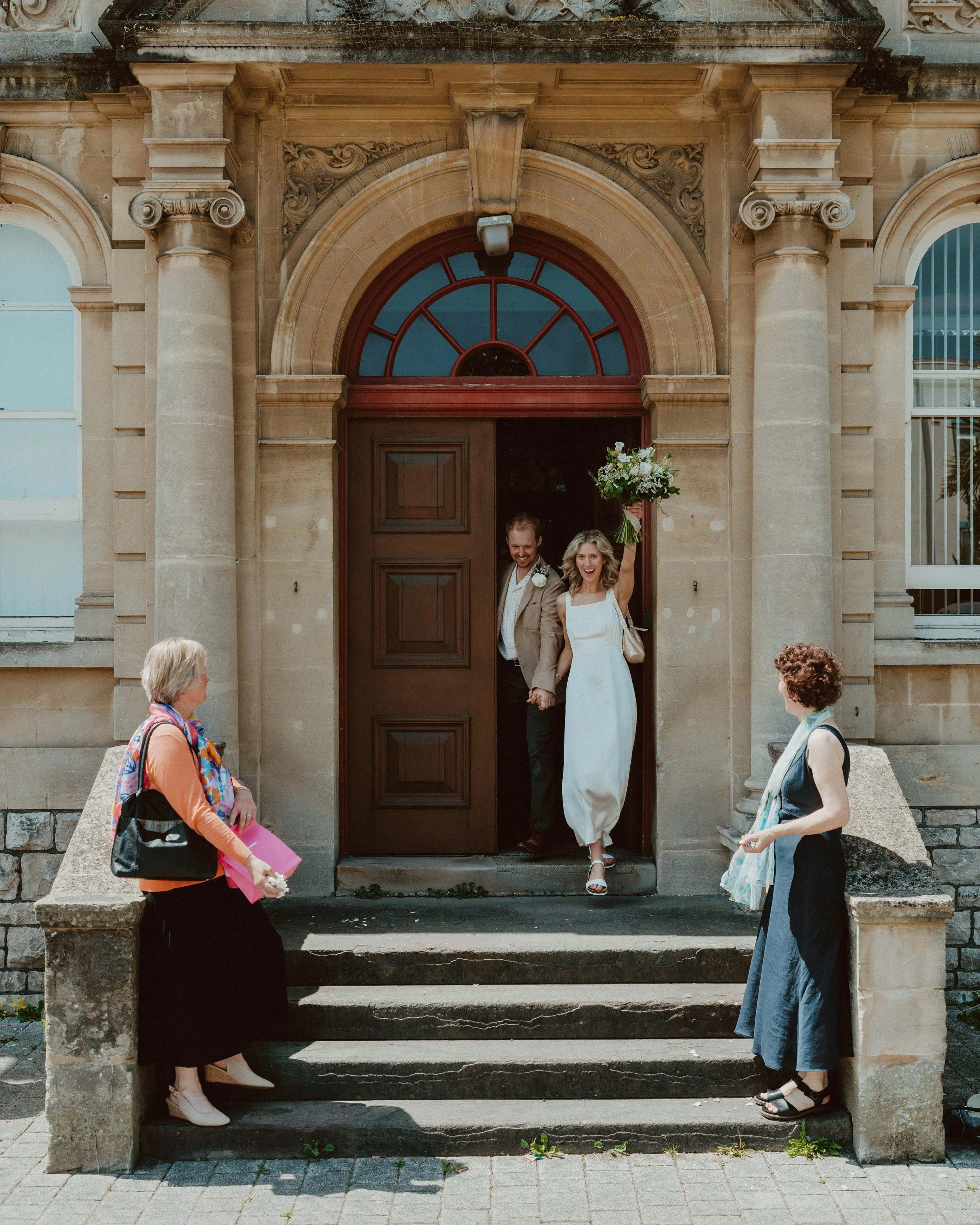A newly married couple exiting a church, holding hands, with the bride smiling and raising her bouquet. They are greeted by two women on the steps outside the church, one with a colorful scarf and the other in a dark dress.