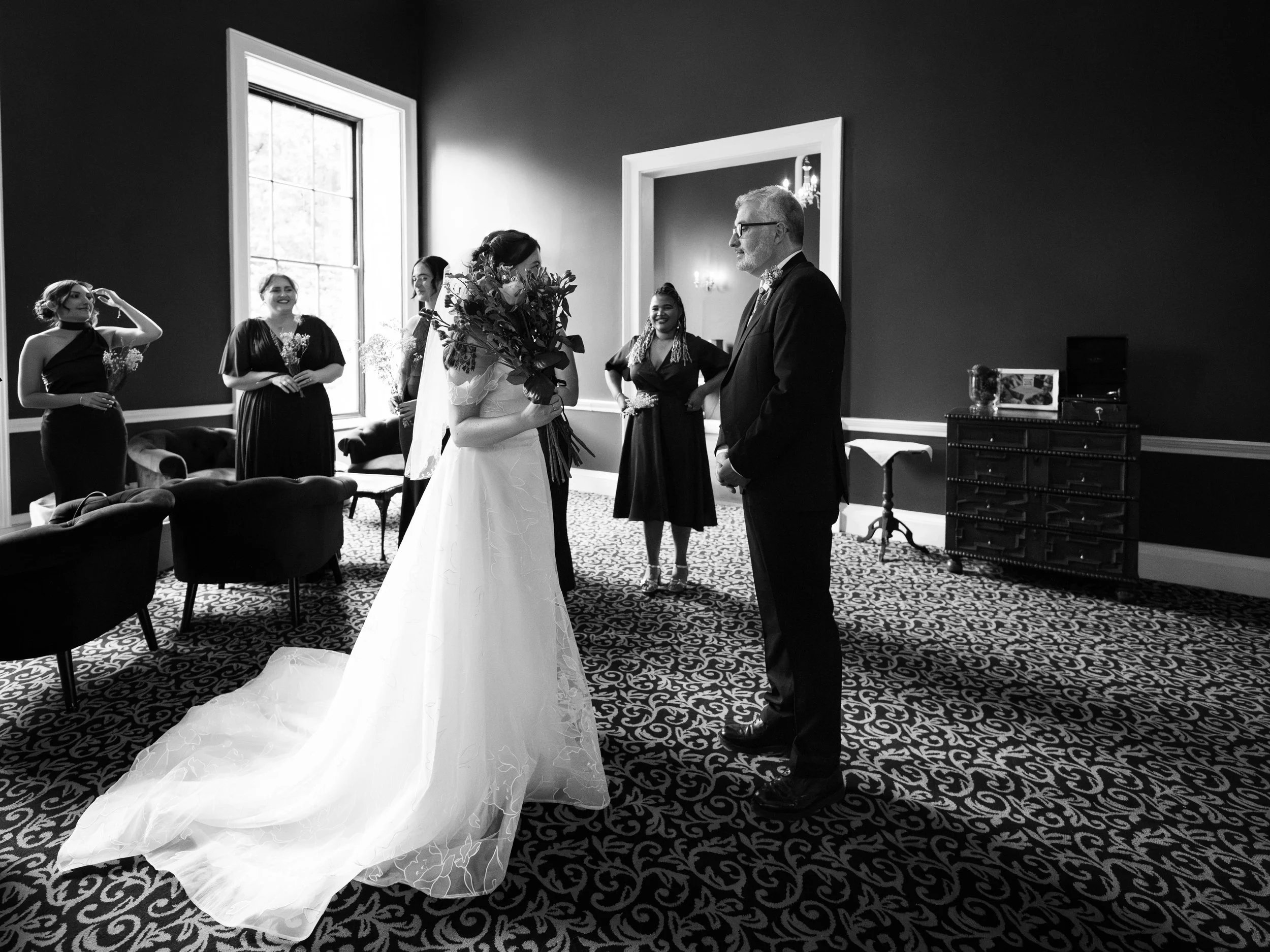 A bride holding a bouquet of flowers in front of a groom during a wedding ceremony, with bridesmaids in black dresses watching in a decorated room with a window and patterned carpet.