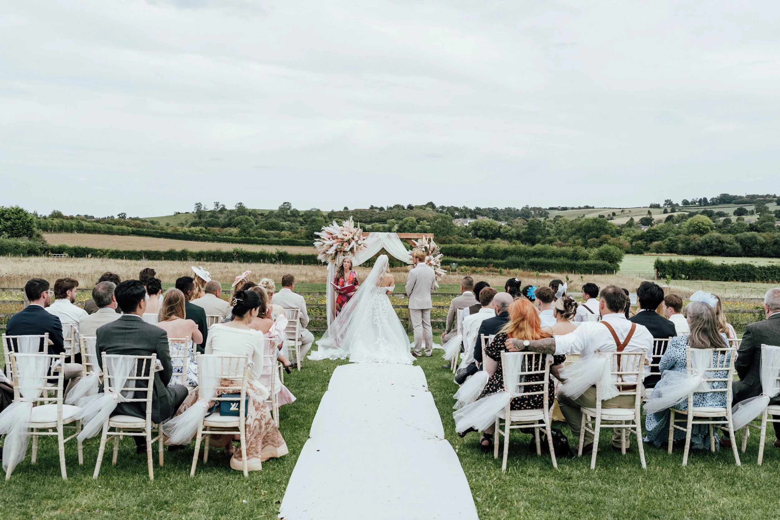 Outdoor wedding ceremony with a bride and groom standing at the altar, surrounded by seated guests on green grass with scenic countryside in the background.