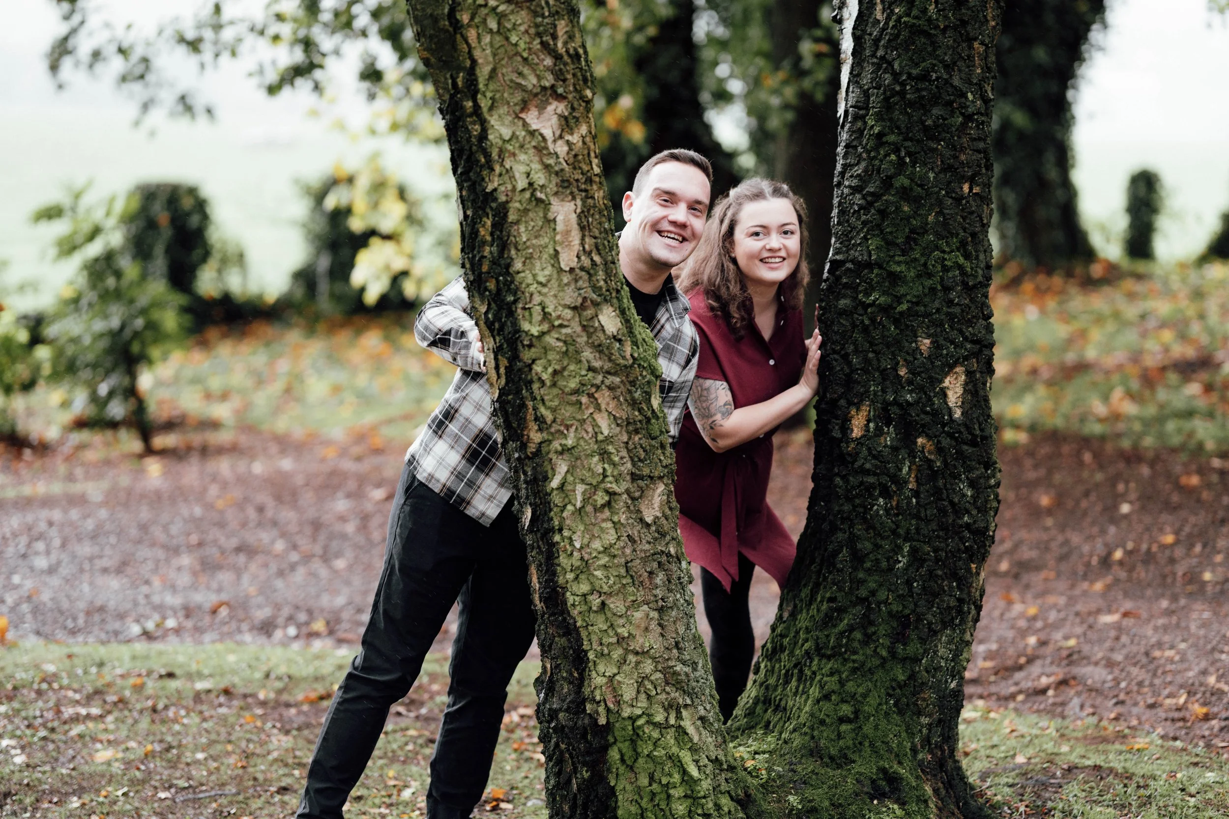 A man and woman peek out from behind trees in a park, smiling at the camera. The man is wearing a plaid shirt and black pants, the woman is wearing a maroon dress with tattoos on her arm. The background features fallen leaves and greenery.