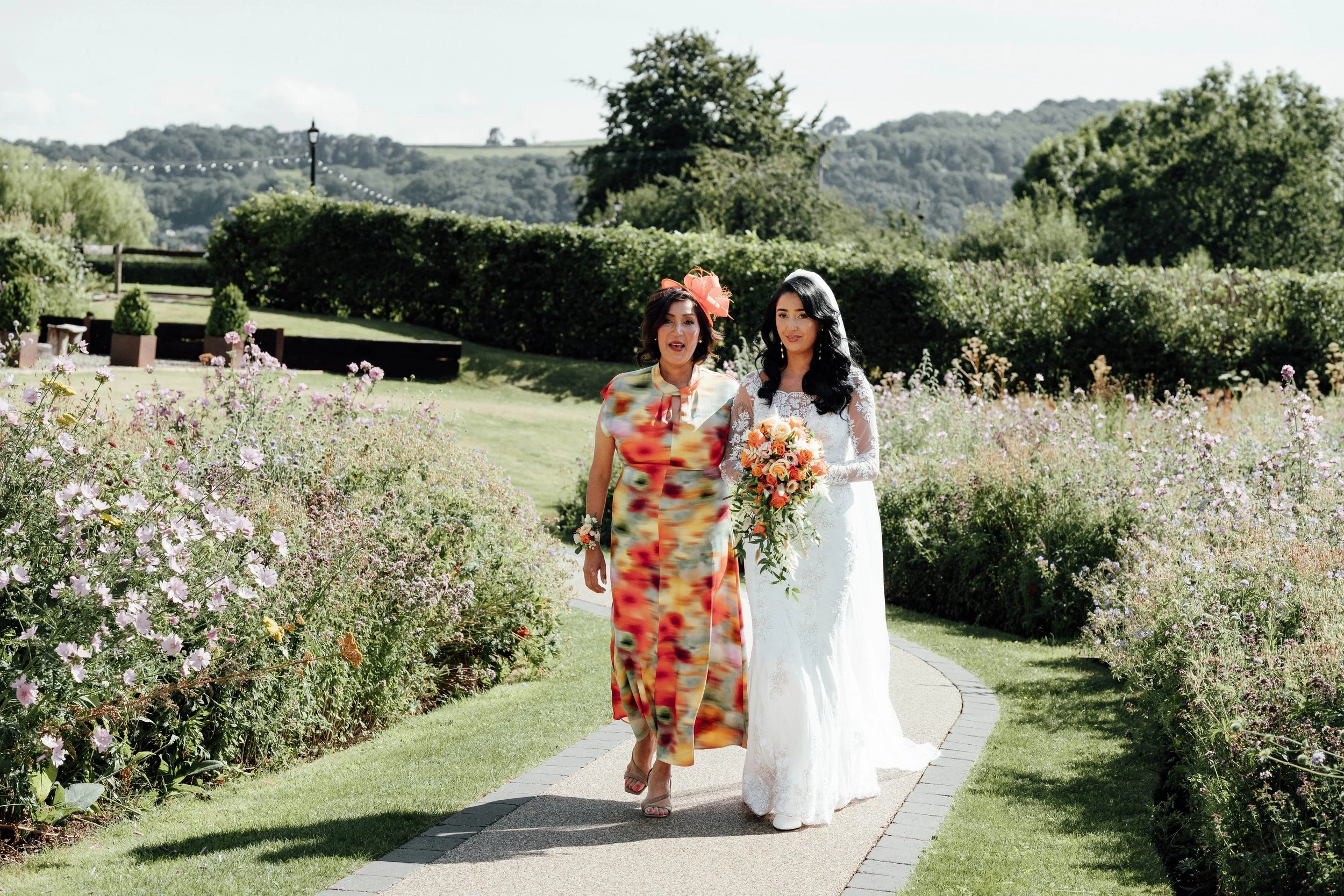 A bride in a white lace wedding dress holding a bouquet walking on a garden path with a woman in a colorful dress and large hat in a scenic outdoor garden setting with greenery and hill in the background.