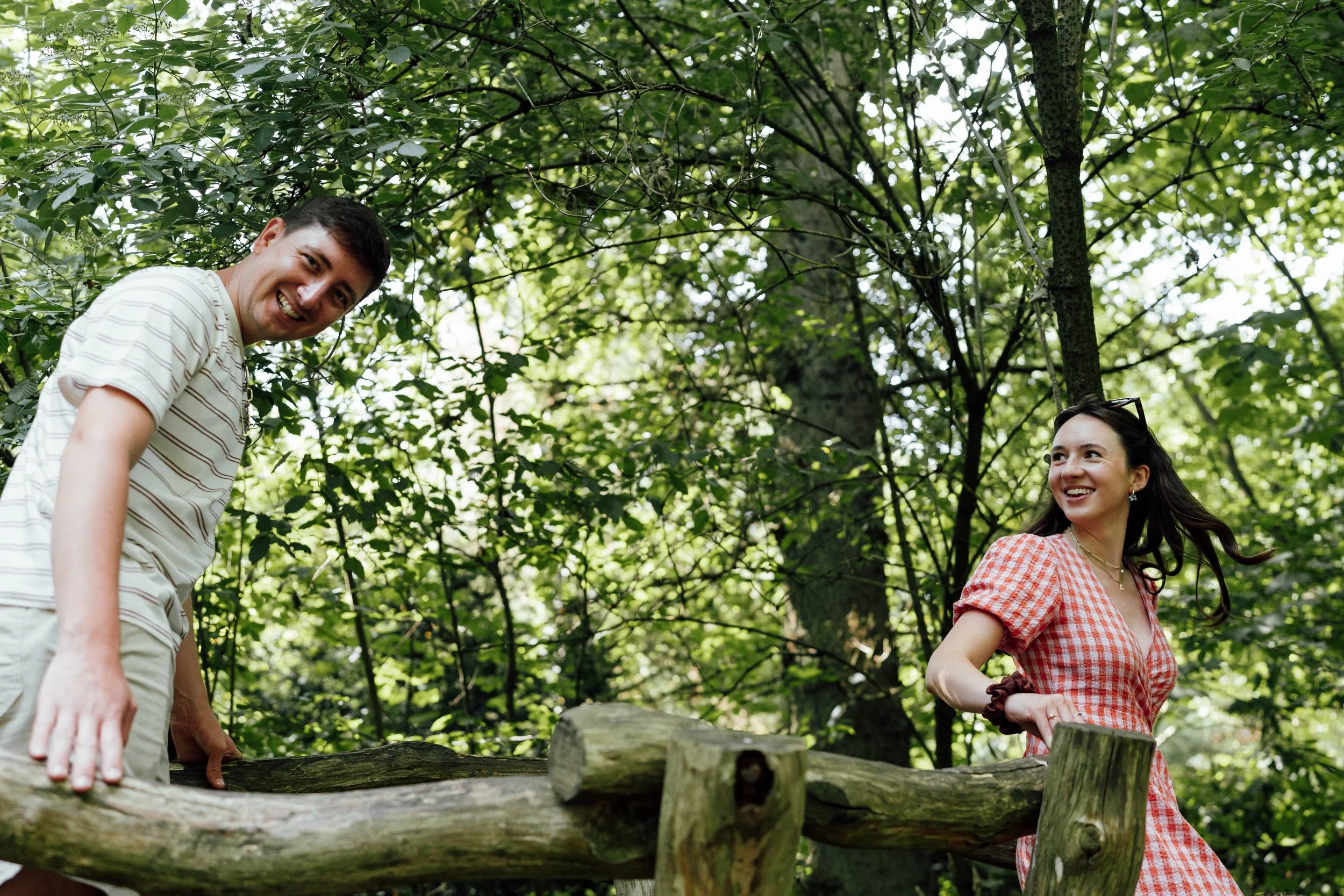 Two smiling young adults, a man and a woman, enjoying time outdoors on a wooden balance beam in a forest surrounded by green trees.