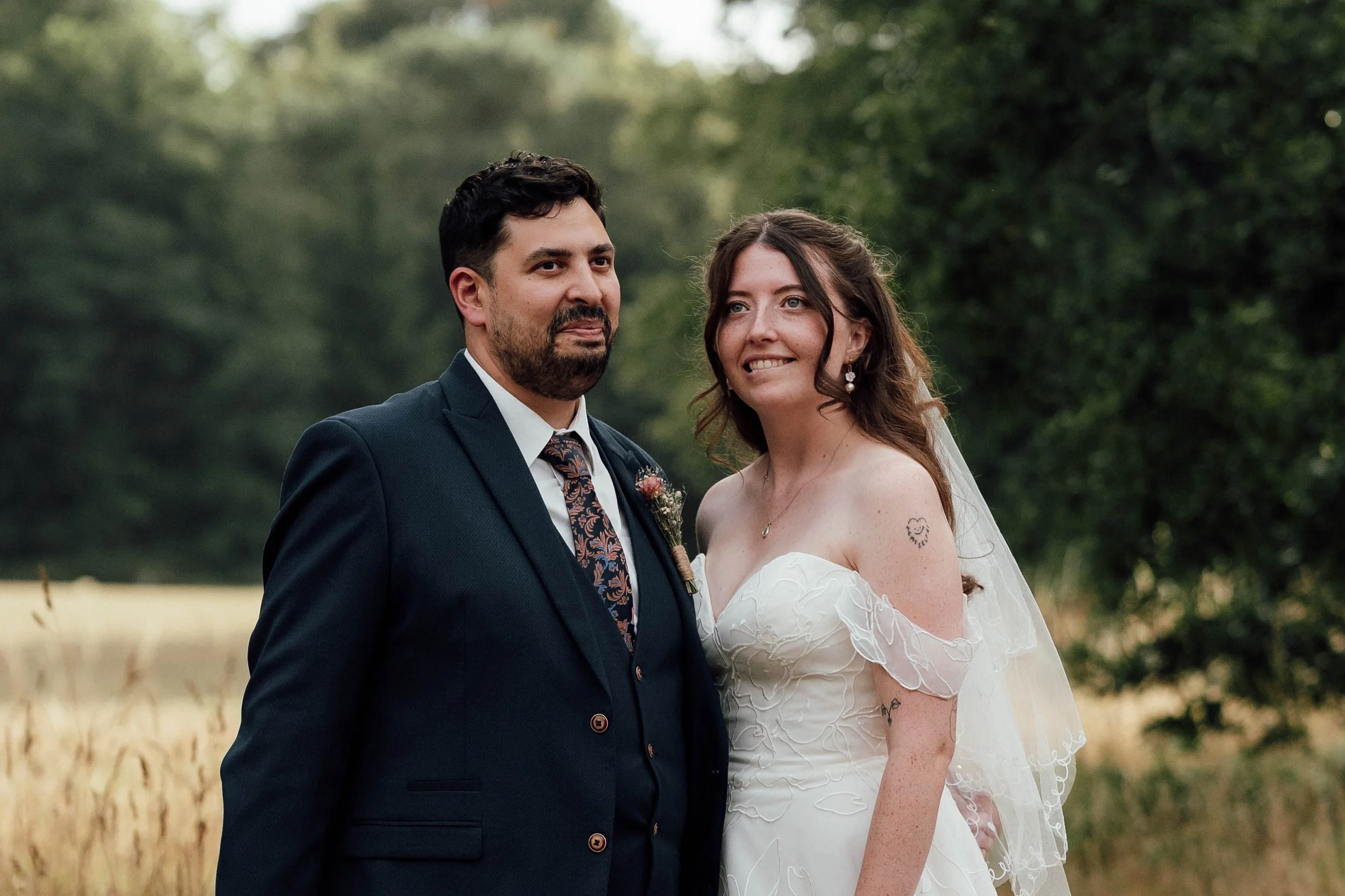 A bride and groom standing outdoors in a field with trees in the background, dressed in wedding attire, smiling softly.