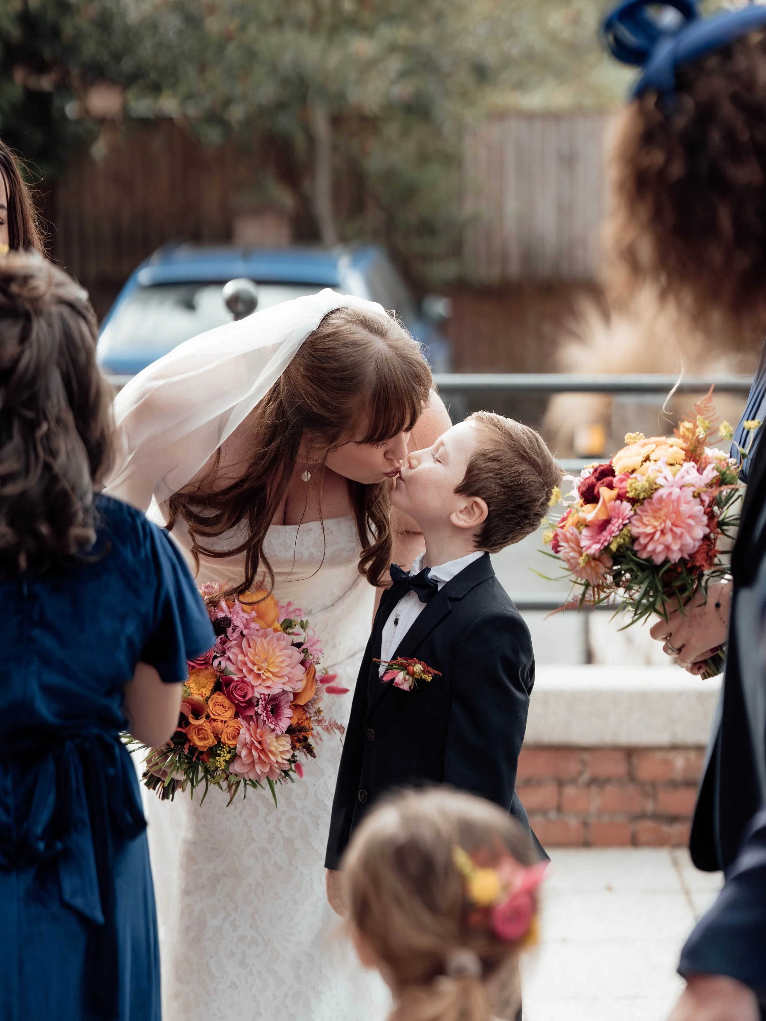 A bride and a young boy are about to kiss during a wedding ceremony, surrounded by bridesmaids and flower girls holding bouquets of colorful flowers, outdoors with trees and a brick wall in the background.