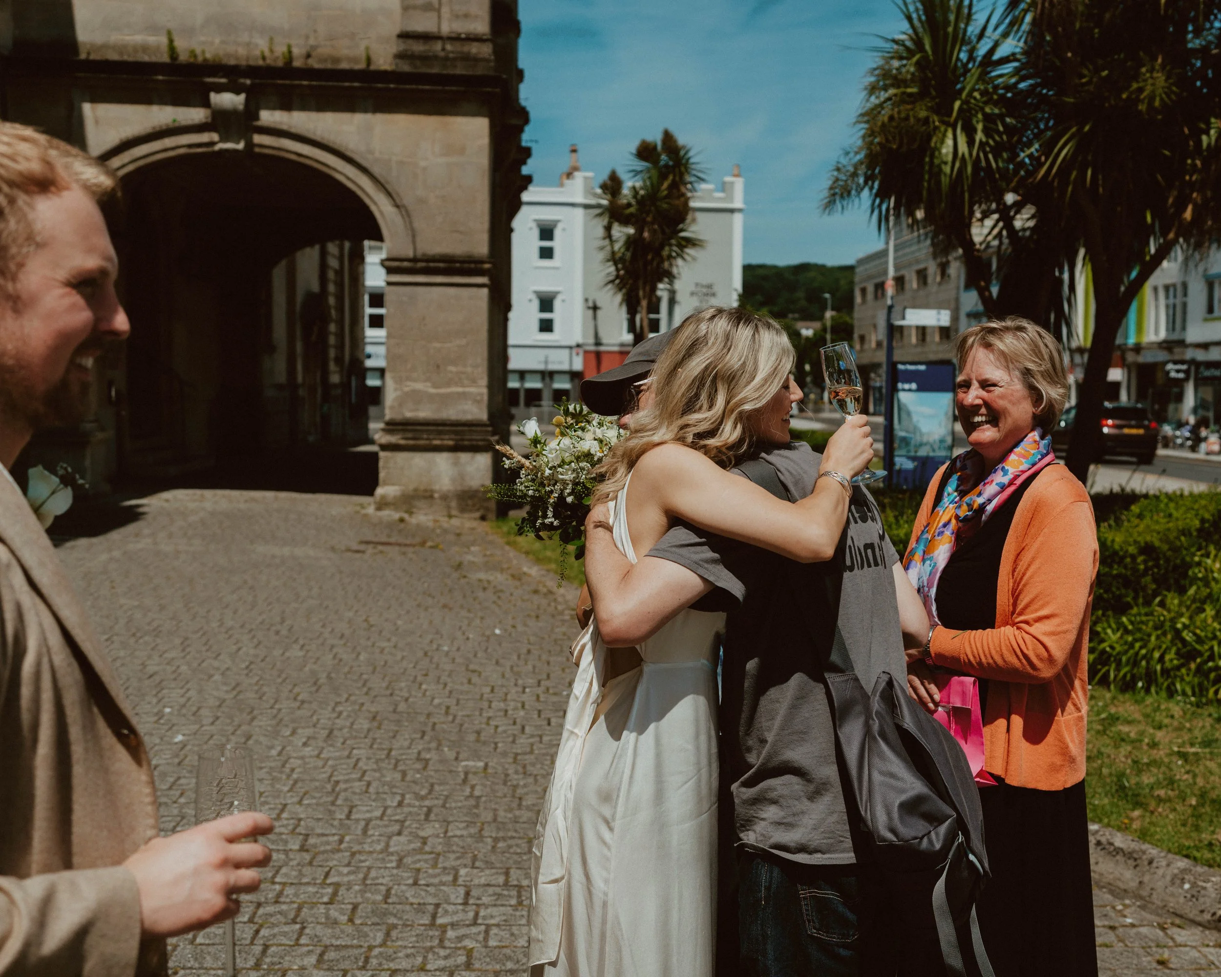 People celebrating outdoors, with a woman in white receiving a hug and holding a glass of champagne, surrounded by other smiling women and men, in front of an archway and trees.