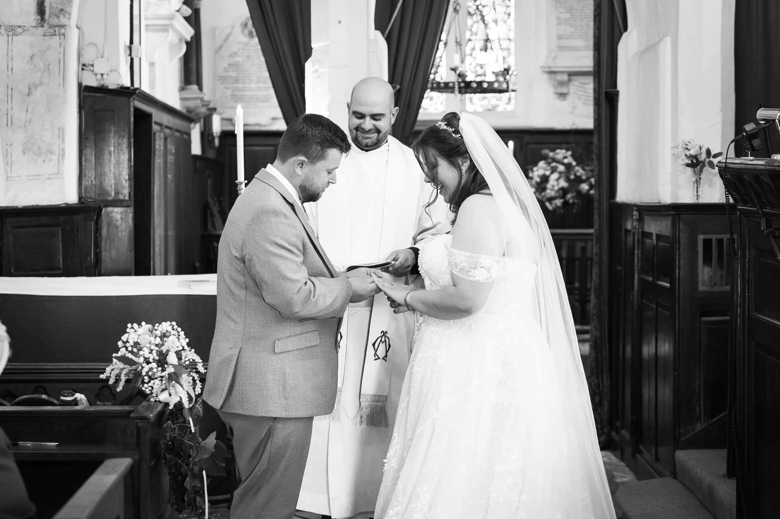 A bride and groom exchanging wedding rings during a ceremony in a church, with a priest officiating, in black and white.