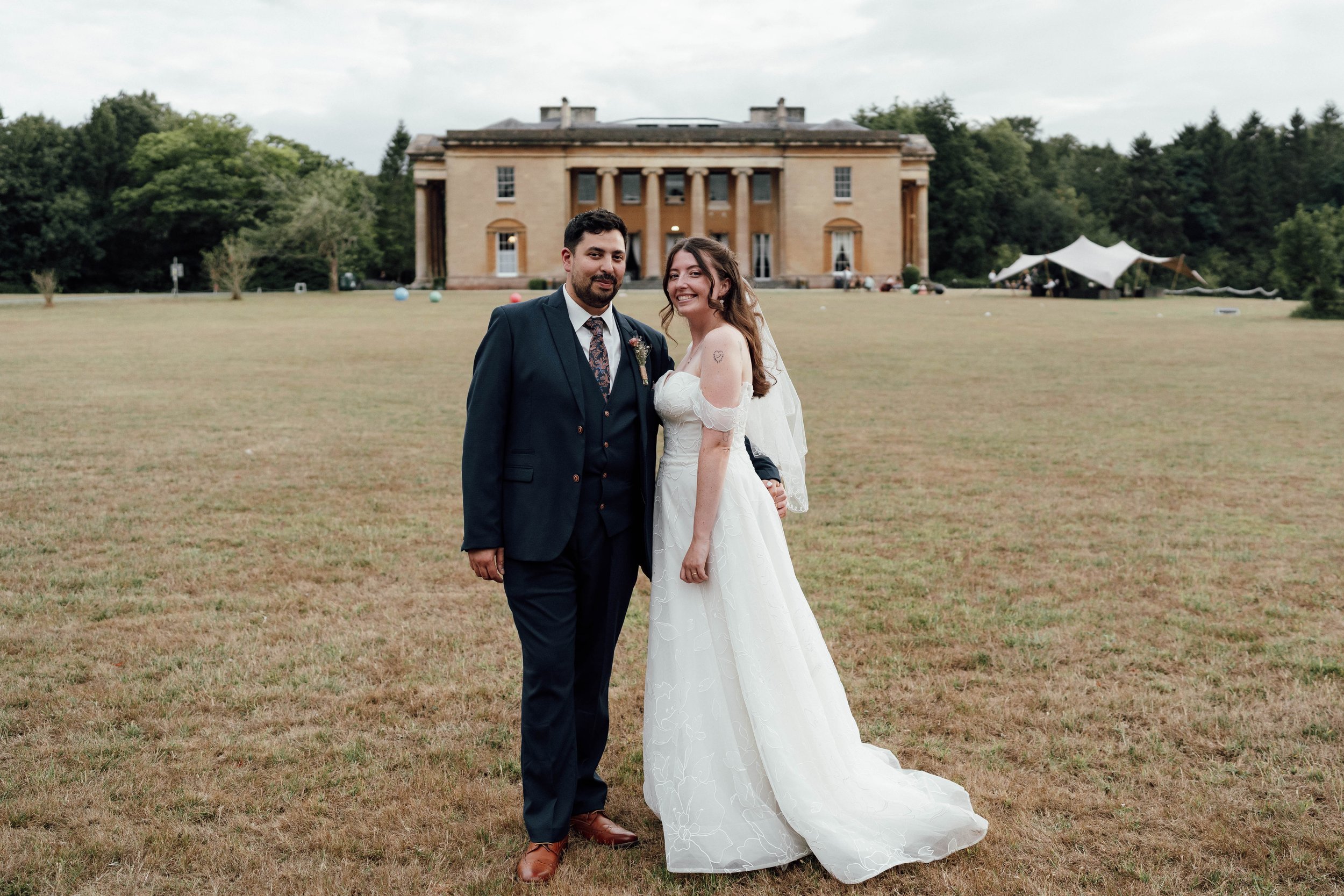 A bride and groom standing on a grassy field in front of a large historic mansion during their wedding. The groom is wearing a dark suit with a patterned tie, and the bride is wearing a white off-the-shoulder wedding dress with a veil. The couple is 