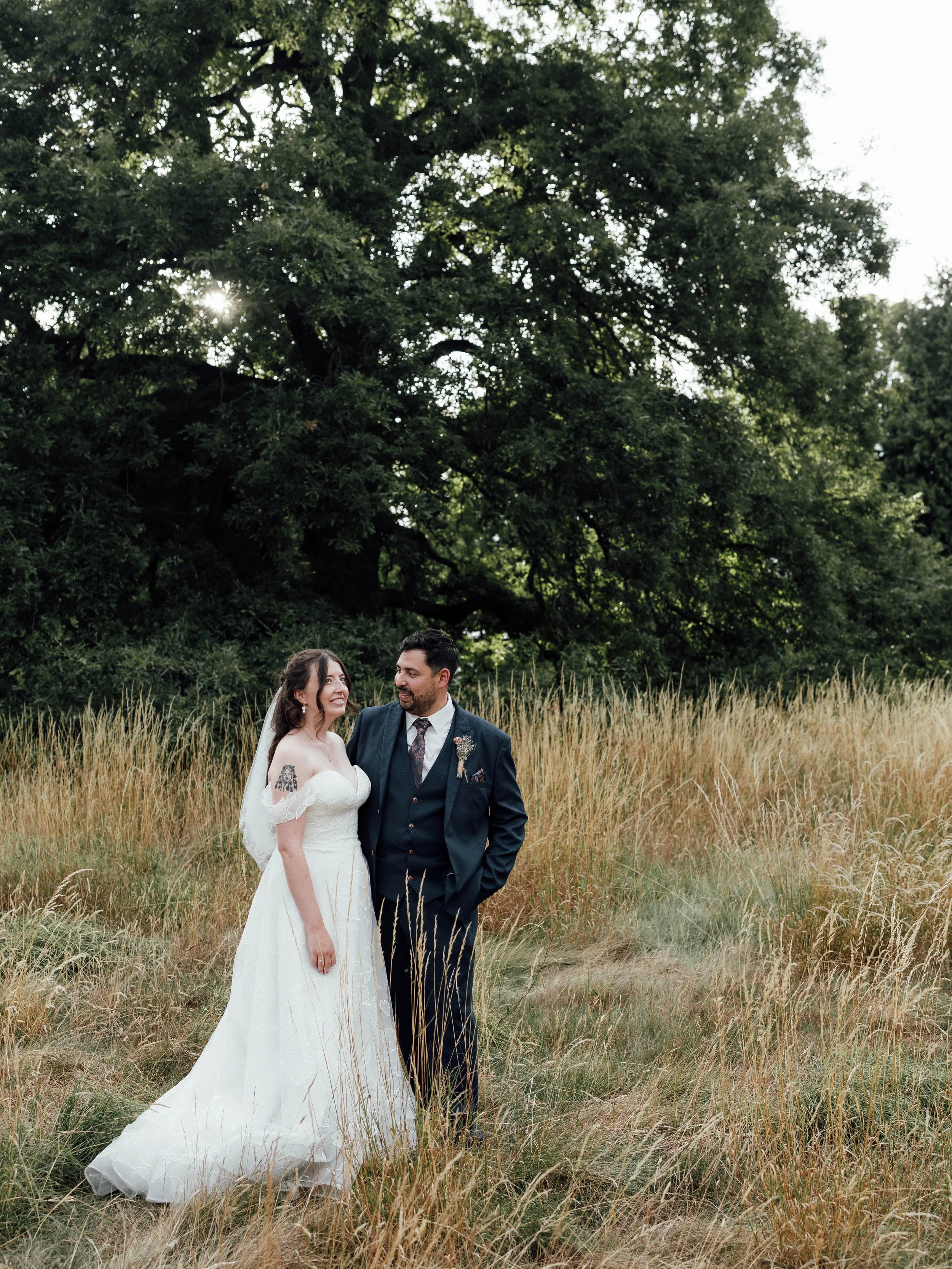 A bride in a white wedding dress and a groom in a dark suit stand in a field of tall grass with a large tree in the background, smiling at each other.