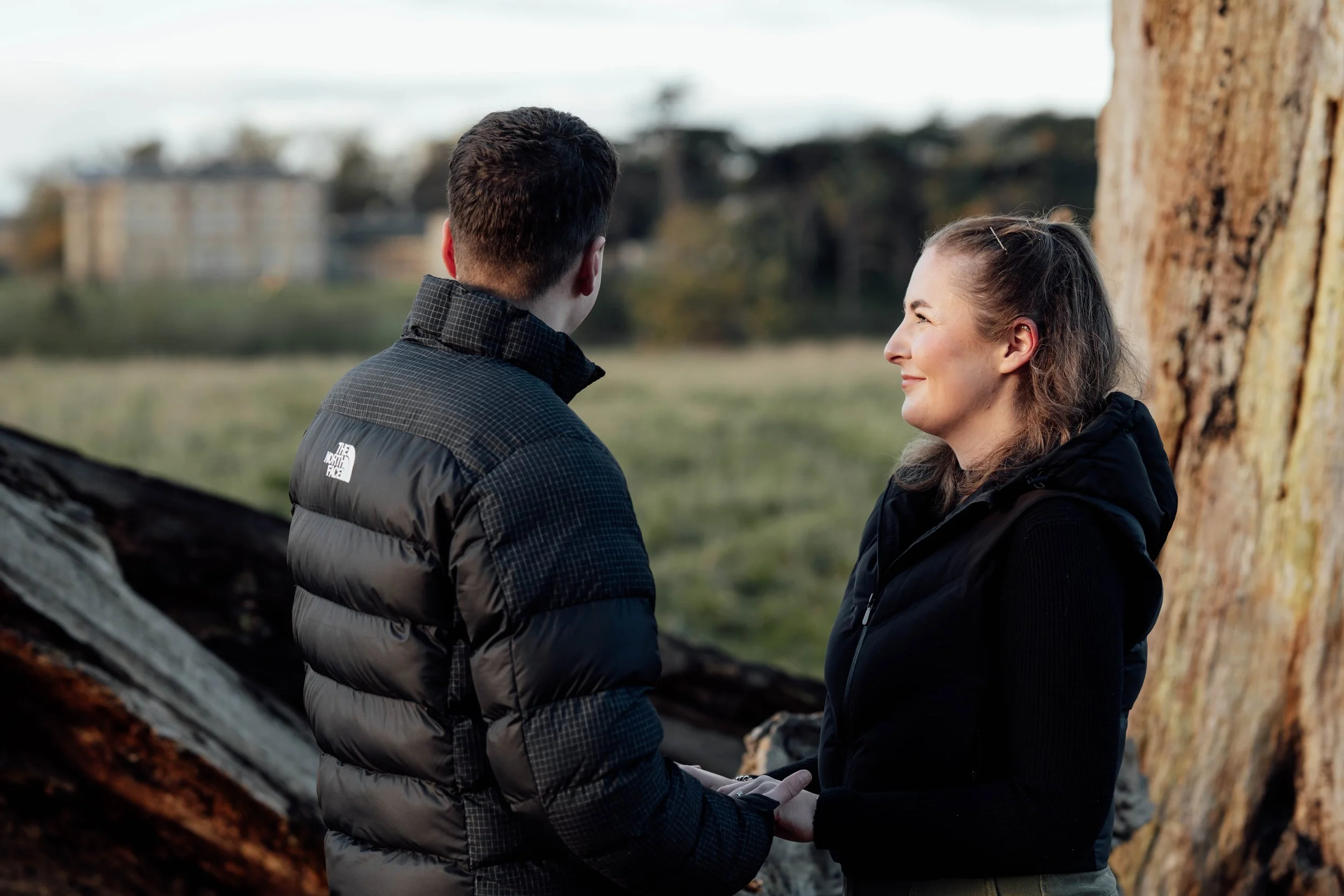 A man and woman holding hands outdoors near a large tree, with buildings in the background.