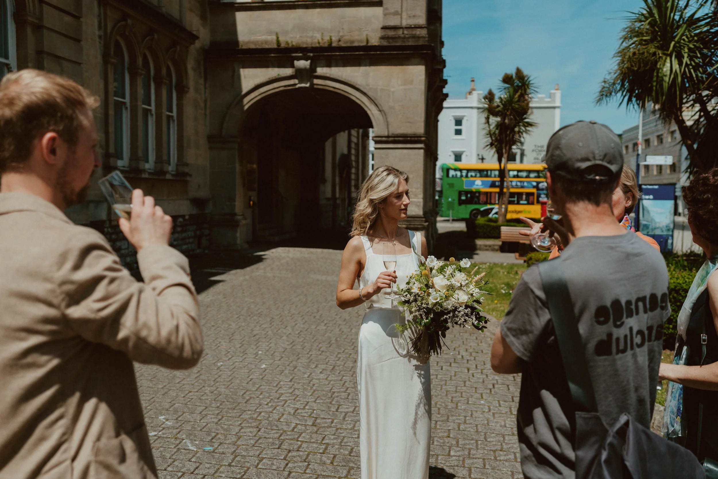 A woman in a white dress holding a bouquet and a glass of wine standing outside while surrounded by people on a sunny day.