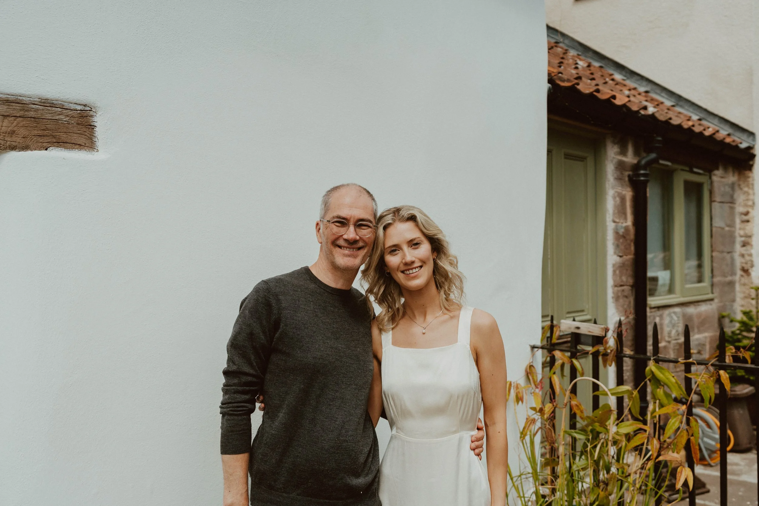 A man and woman smiling outdoors in front of a white wall near a house with brick and green window frames.