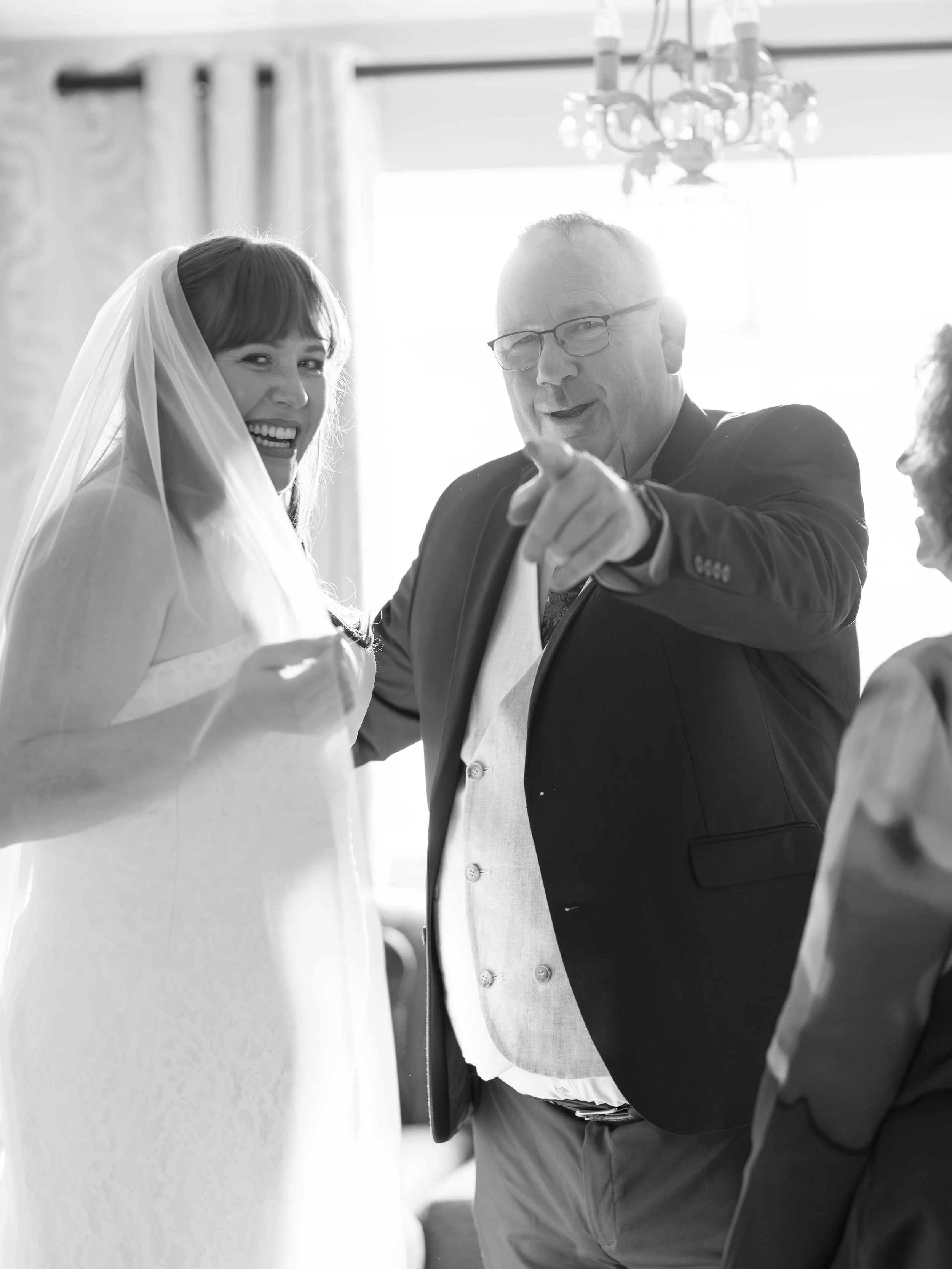 Black and white photo of a bride laughing with a man pointing at the camera, inside a decorated room with large window and chandelier.