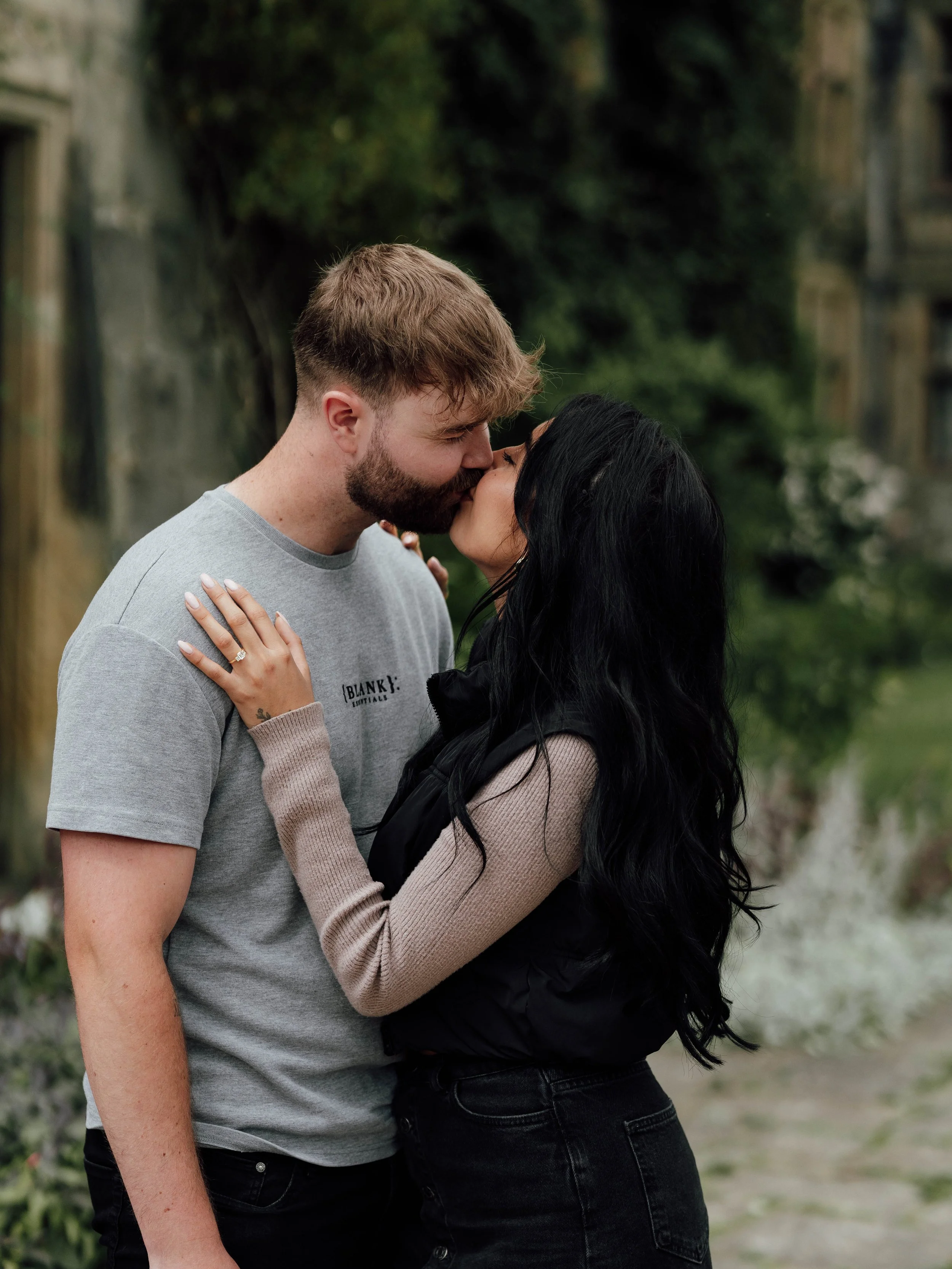 A couple sharing a kiss outdoors with trees and buildings in the background.