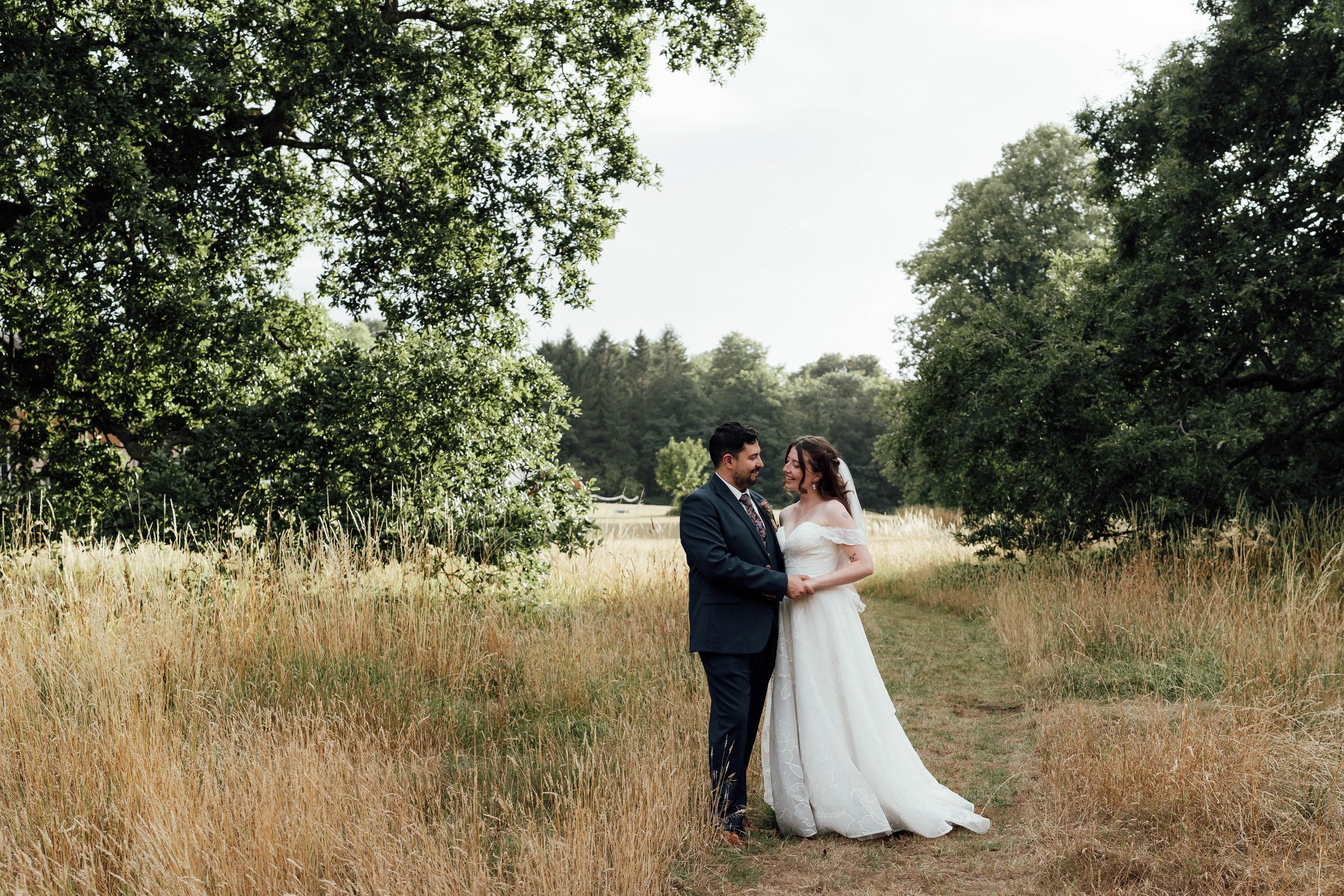 A bride and groom standing on a grassy path amid tall grasses and trees, holding hands and smiling at each other, outdoors on a sunny day.