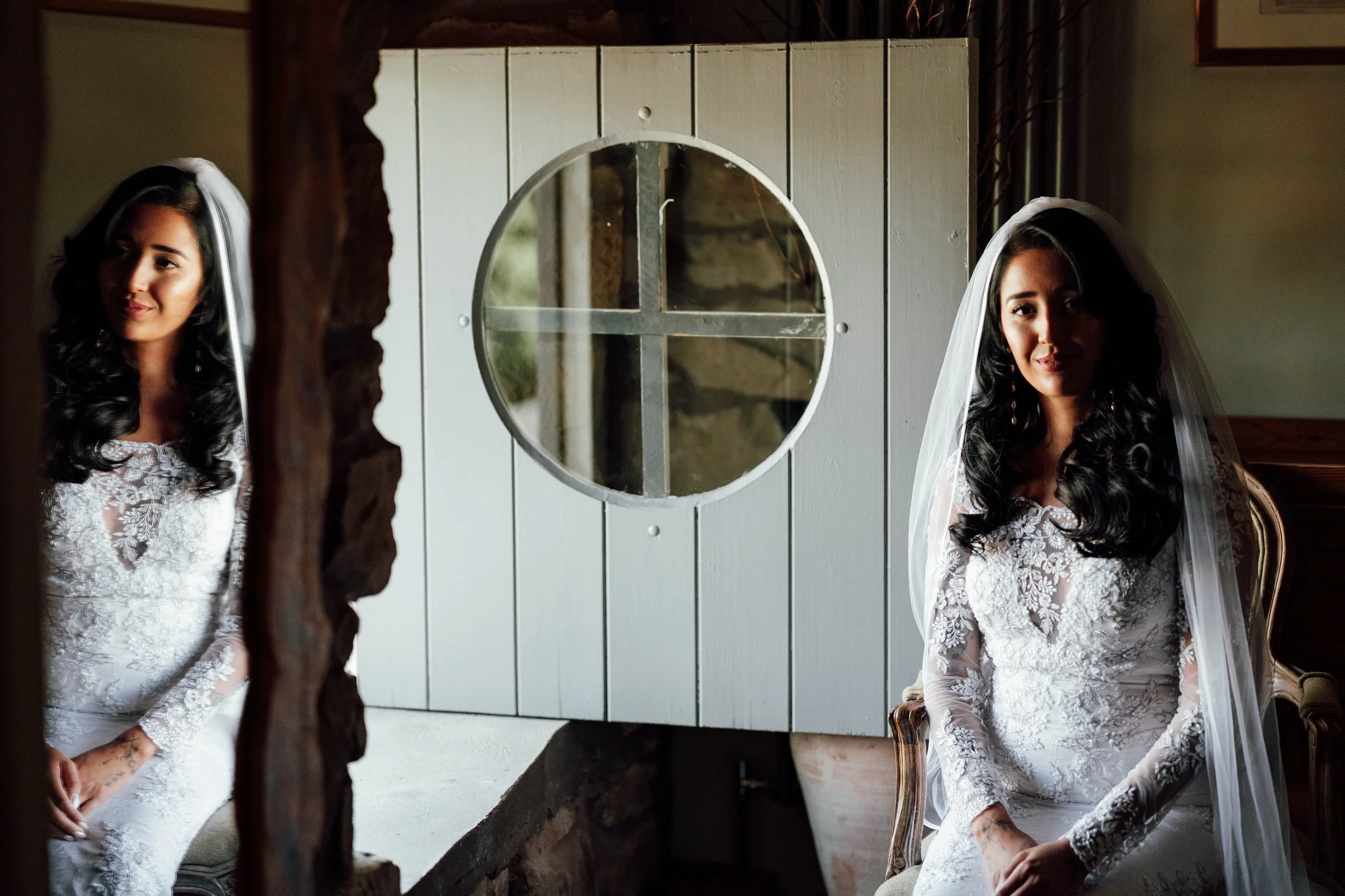A bride with long dark curly hair in a white lace wedding dress and veil, sitting on a vintage wooden chair near a mirror showing her reflection, in a rustic room with wooden and stone accents.
