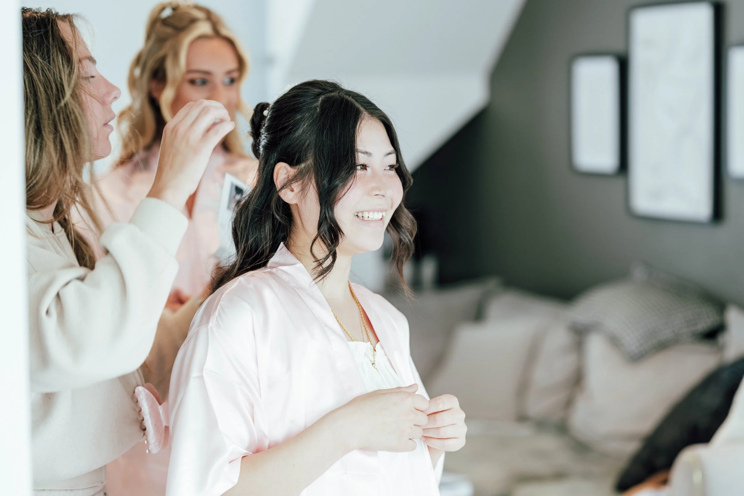 A woman with dark wavy hair smiling as two women style her hair, in a room with framed pictures on the wall.