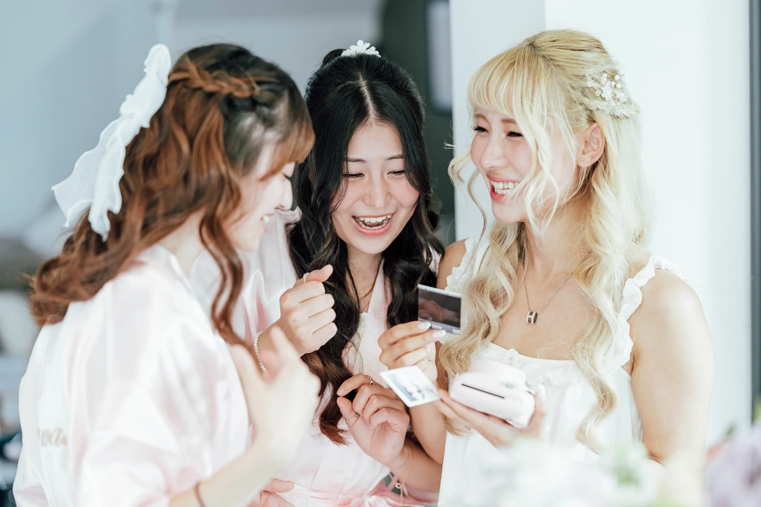 Three women in white dresses look at a photo or note together, smiling and sharing a joyful moment.