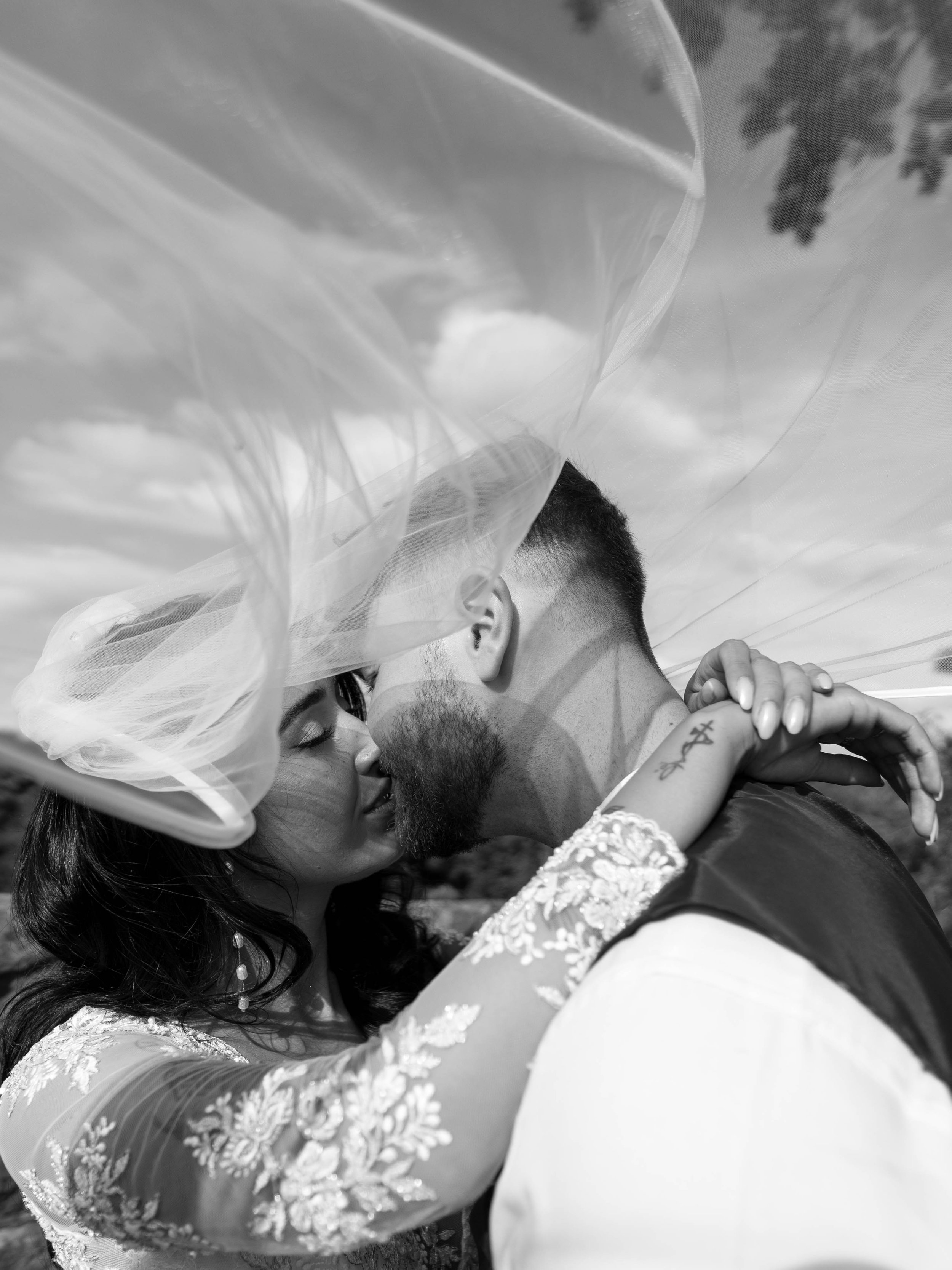 Black and white photo of a couple kissing, with a veil covering their faces, outdoor setting, close-up perspective.
