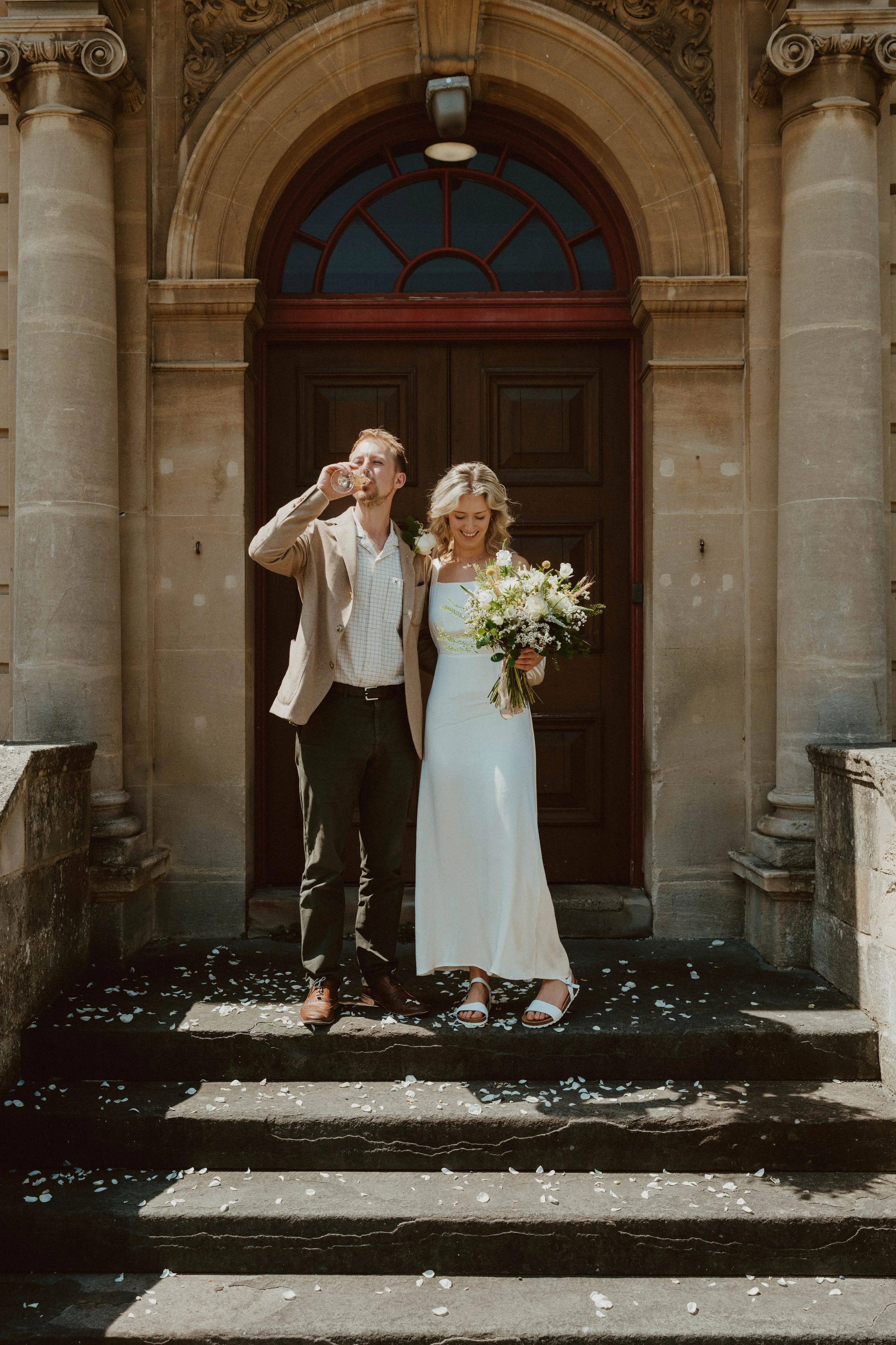 A bride and groom standing on steps outside a church, celebrating, with the bride holding a bouquet and the groom drinking from a glass.