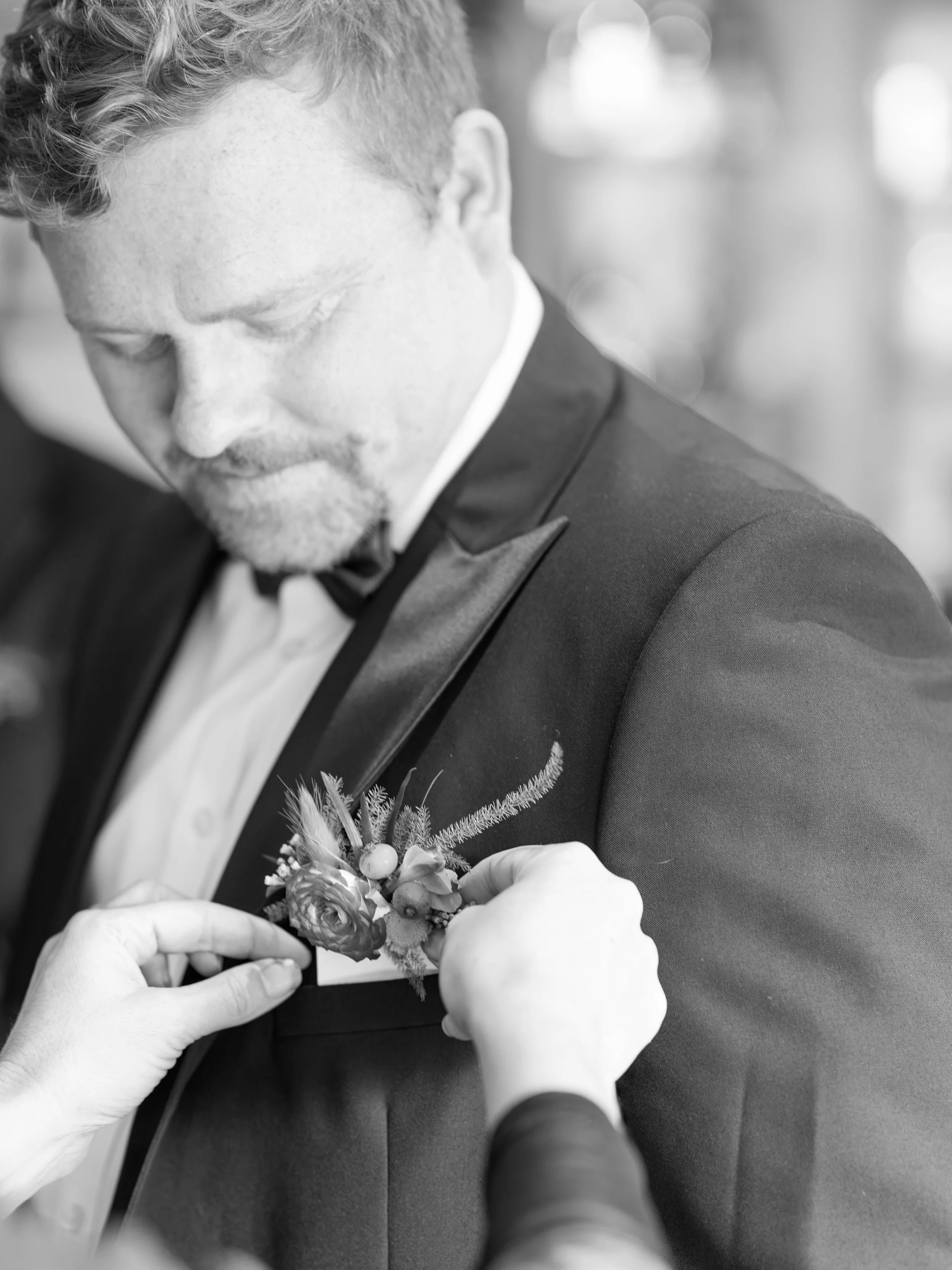 A man in formal attire, likely a suit and bow tie, is being assisted with a boutonniere on his lapel.