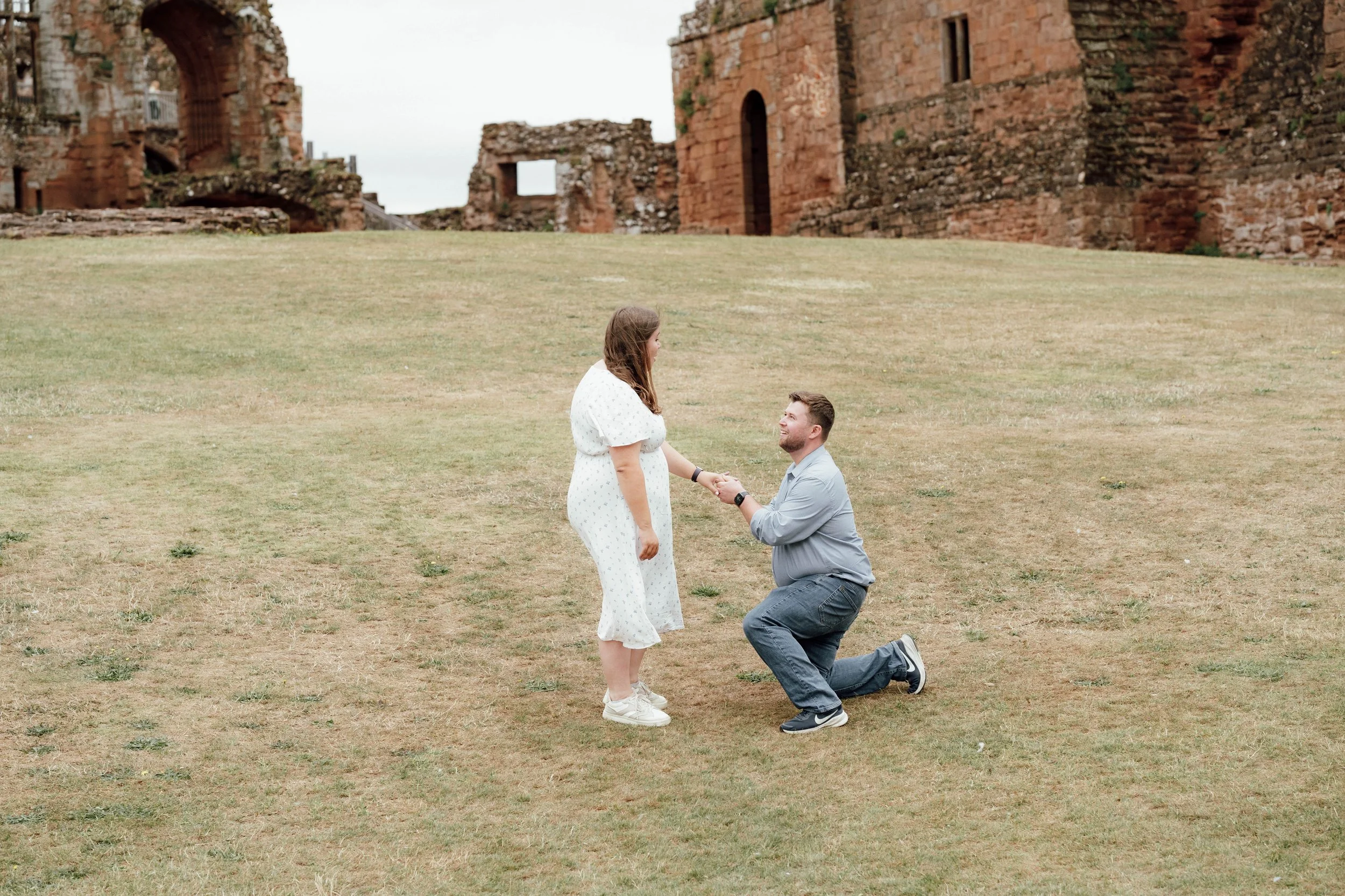 A man on one knee proposing marriage to a woman on a grassy area with old brick ruins in the background.