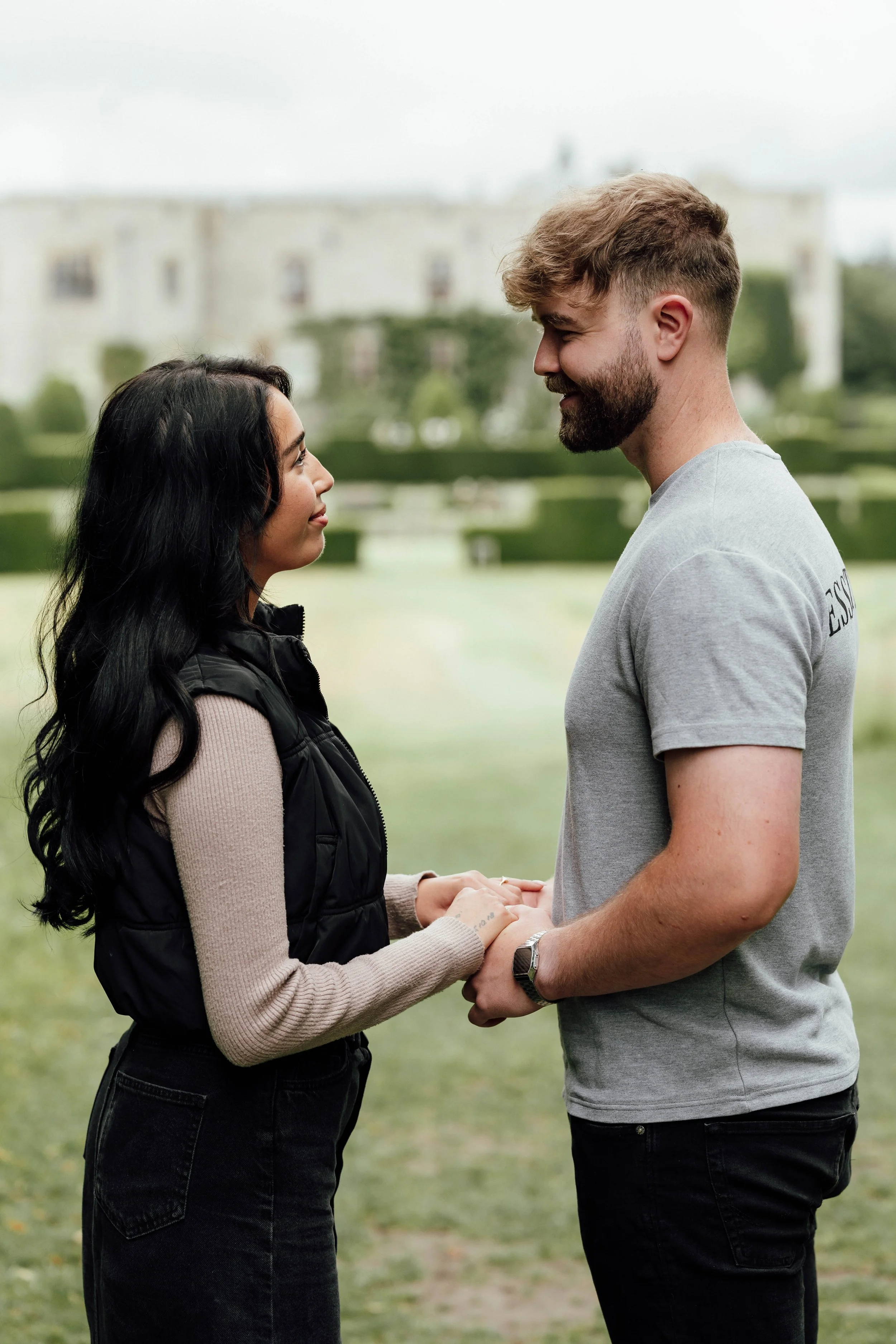A man and woman standing face-to-face outdoors, holding hands and gazing into each other's eyes, with a blurred building in the background.