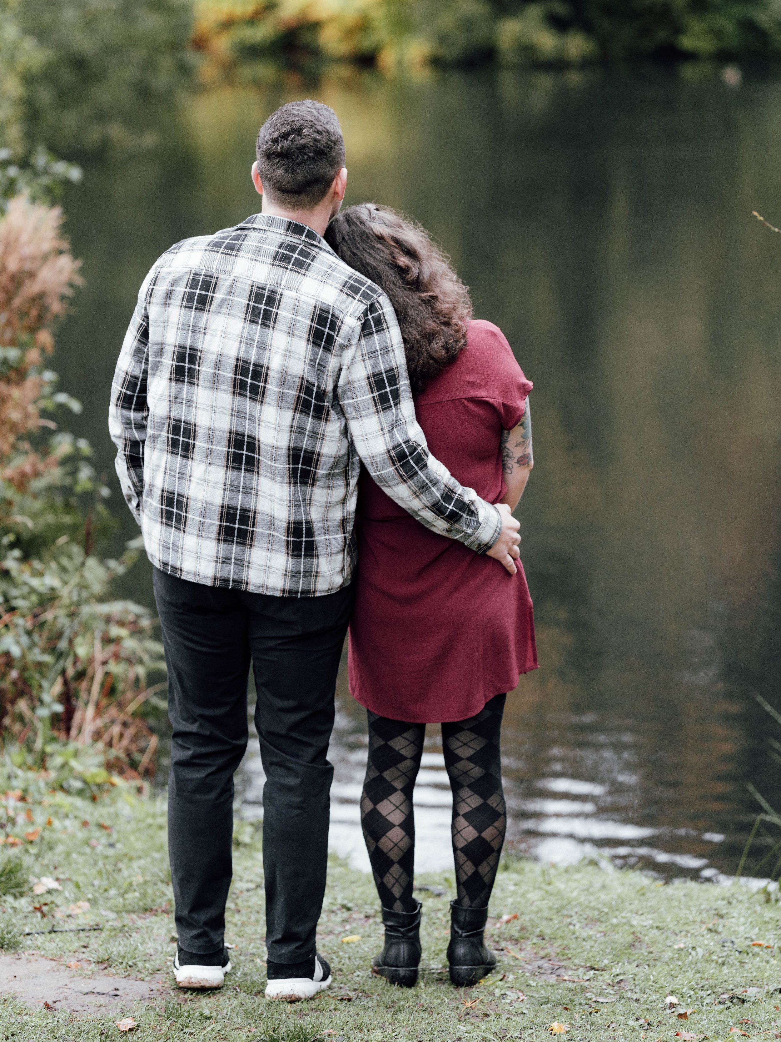 A couple stands close together by a lake, embracing and looking at the water, with trees in the background.