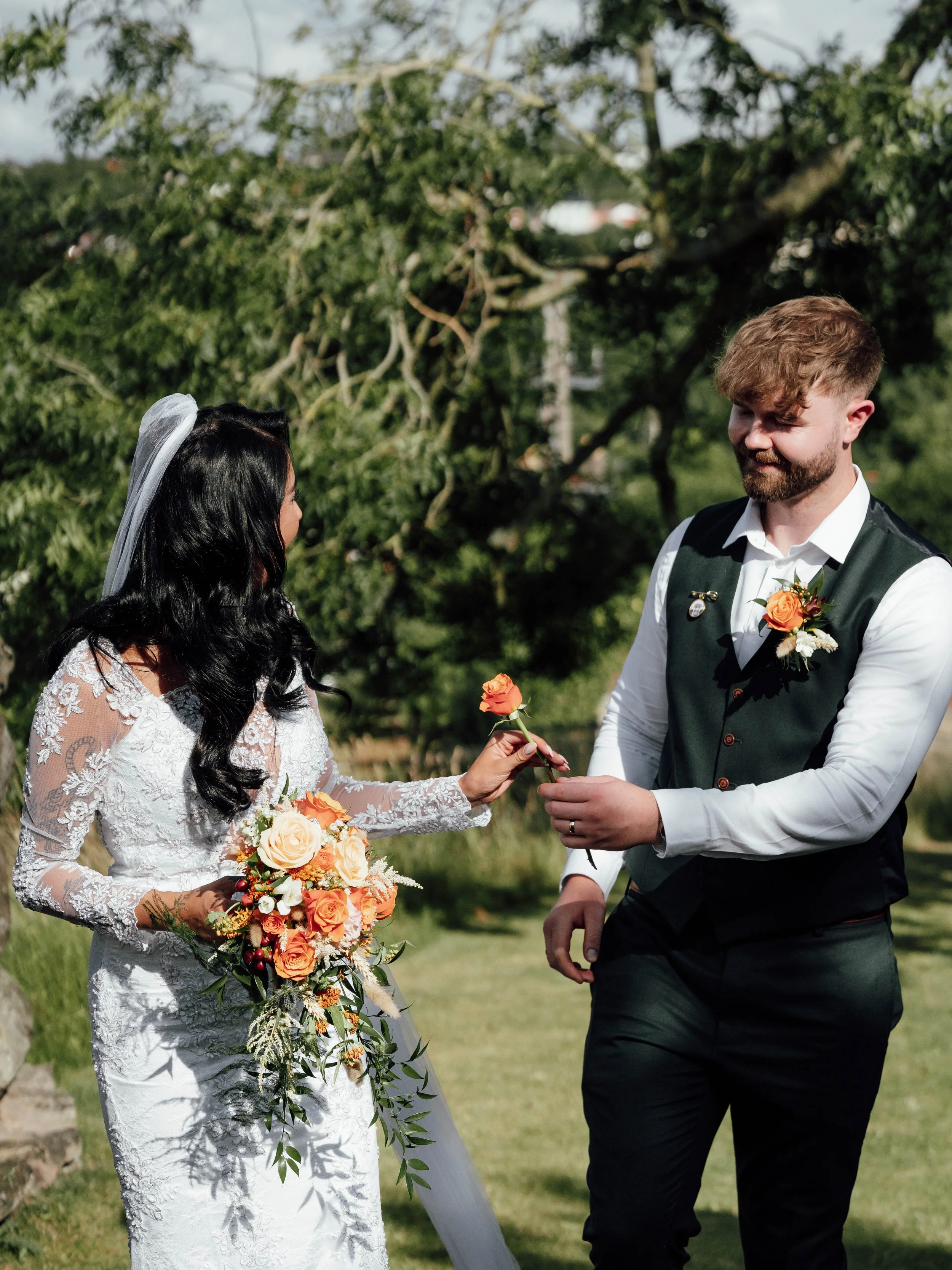 Bride and groom exchanging flowers outdoors during wedding ceremony, with trees and greenery in background.