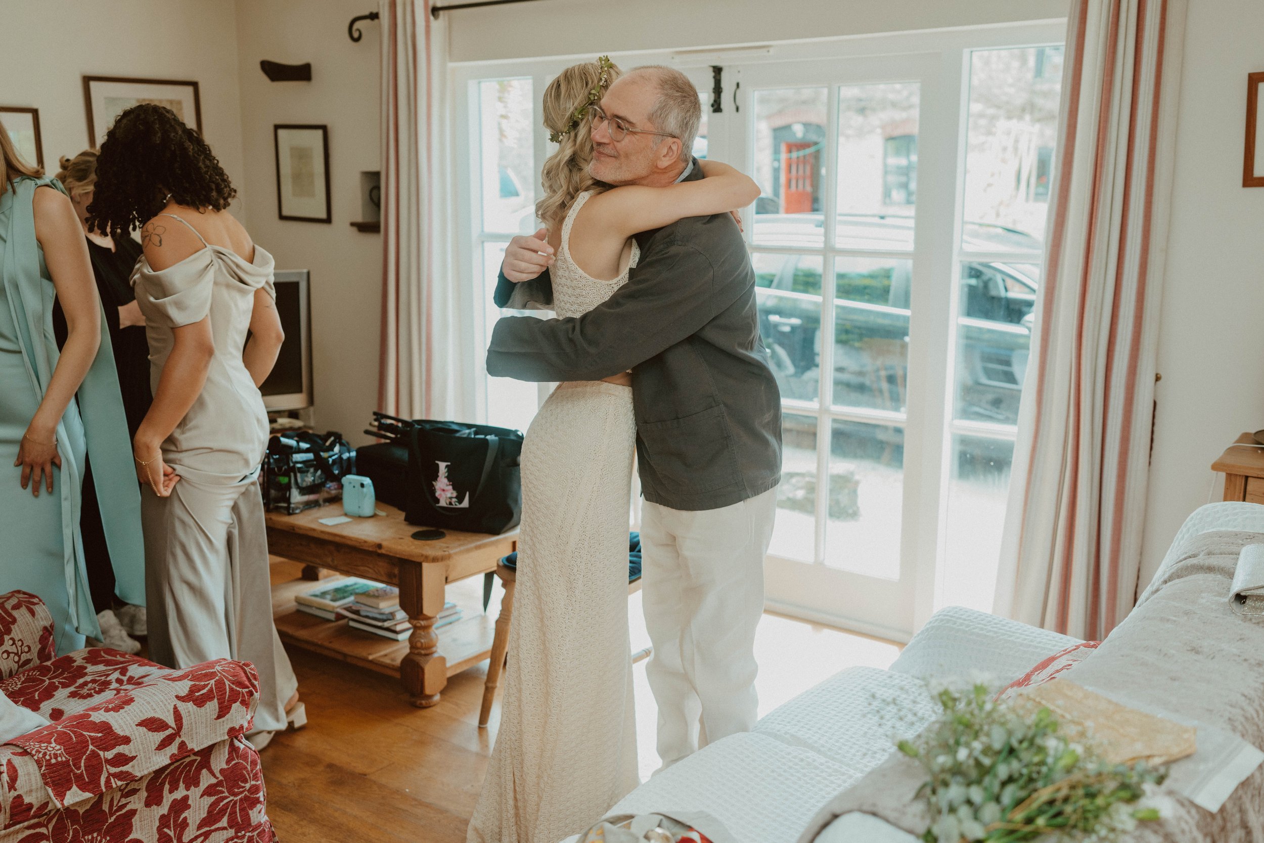 A woman in a white dress hugging an older man in a black jacket inside a living room with a glass door and a window. There are other women in the background preparing for a special occasion.