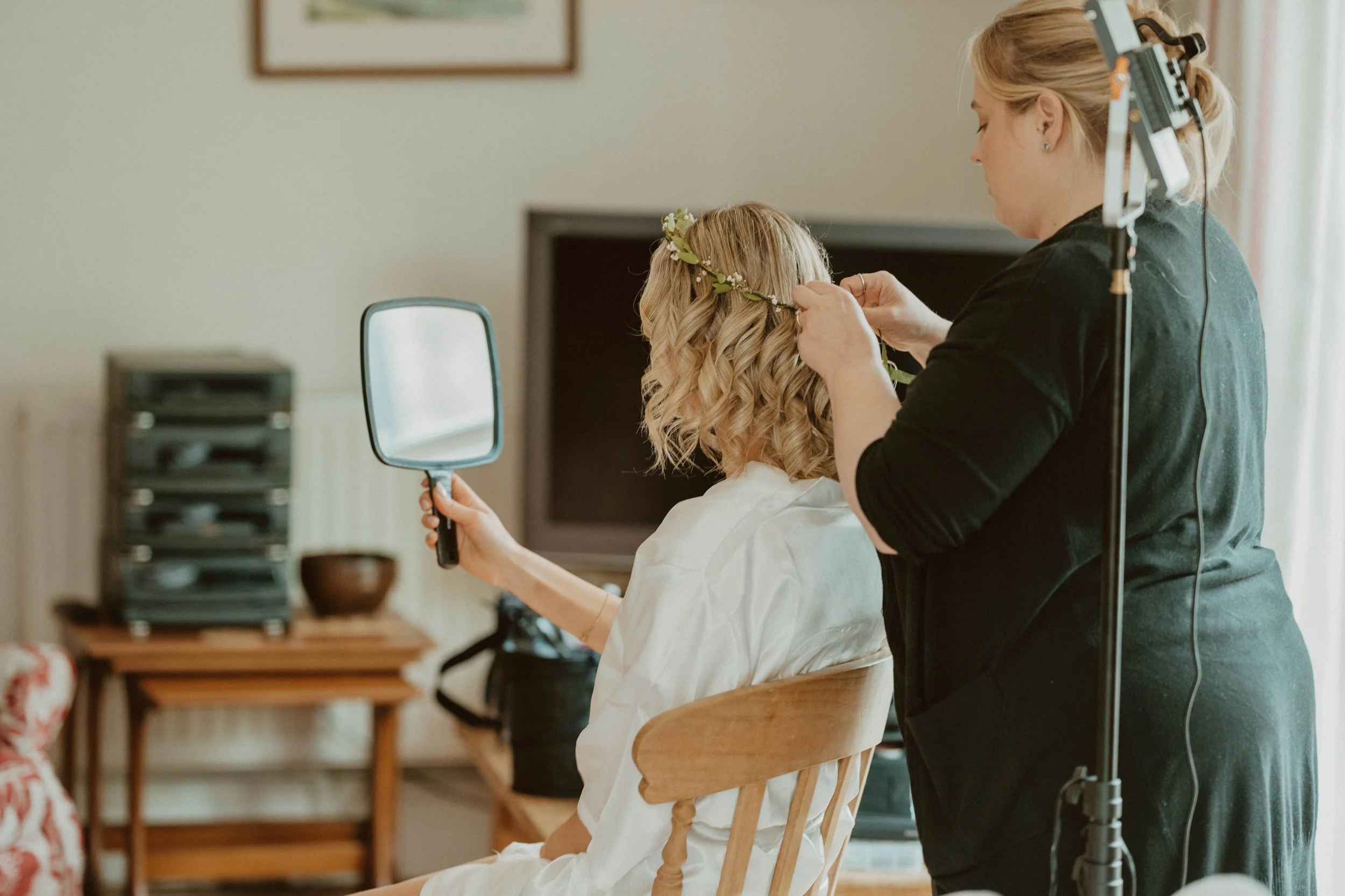 A woman with blonde, curly hair sitting on a wooden chair while a stylist with blonde hair, dressed in black, styles her hair and places a floral crown on her head, with a mirror in hand and studio lights nearby.