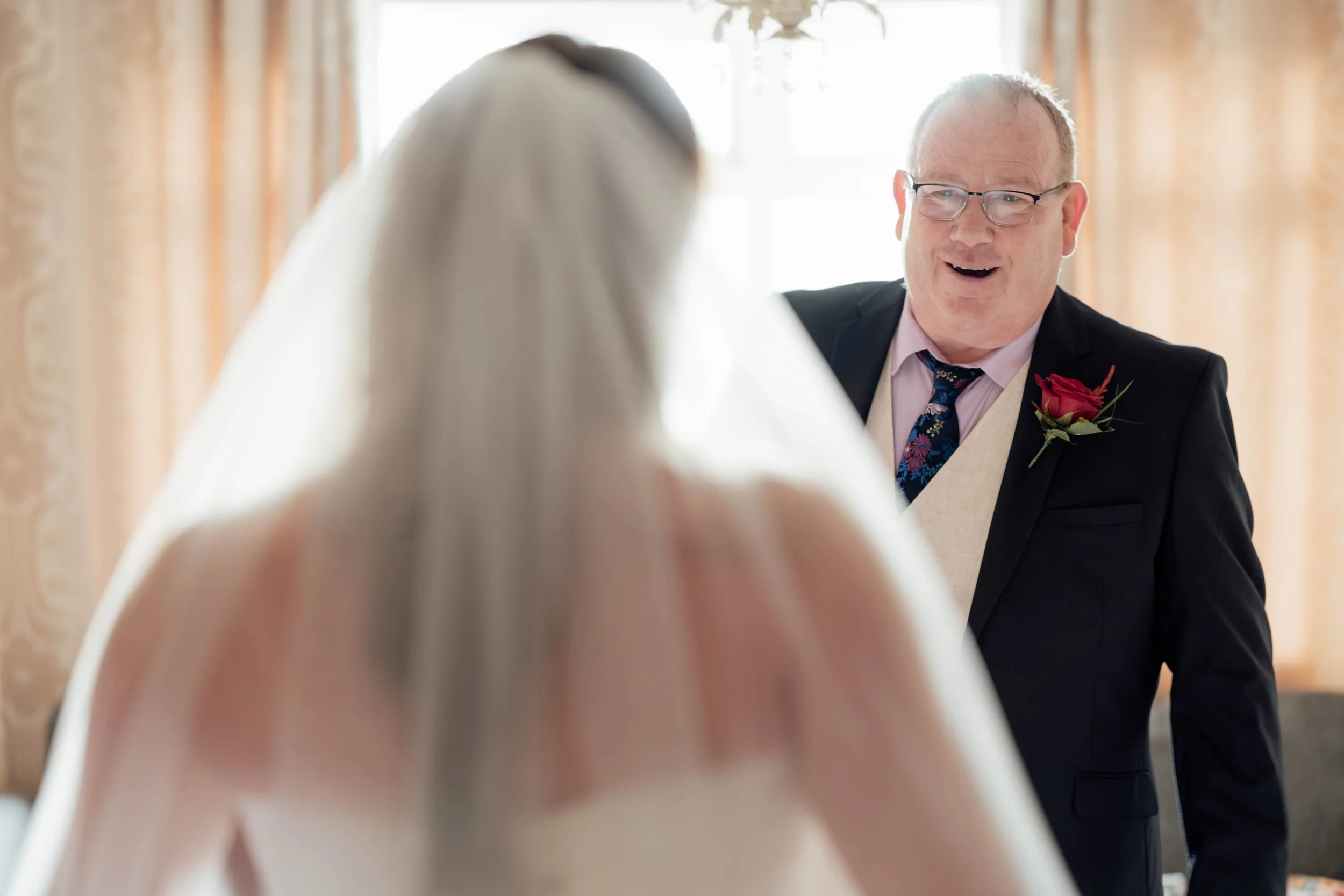 A joyful moment between a groom and an older man, possibly his father or officiant, during a wedding, with warm sunlight and elegant curtains in the background.