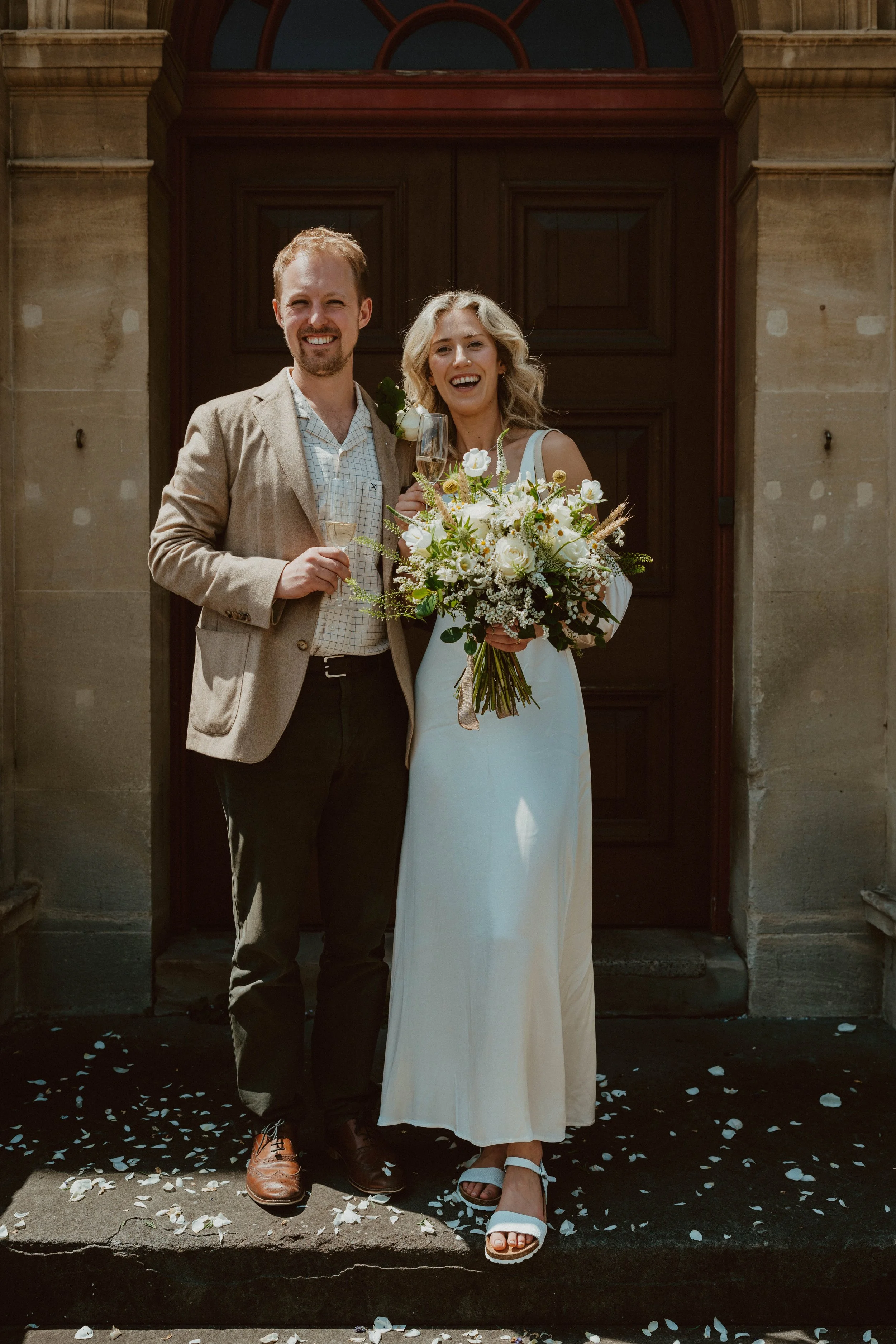 A happy bride and groom stand together outside a church, holding glasses of champagne. The bride is holding a large bouquet of white and green flowers, and they are both smiling.