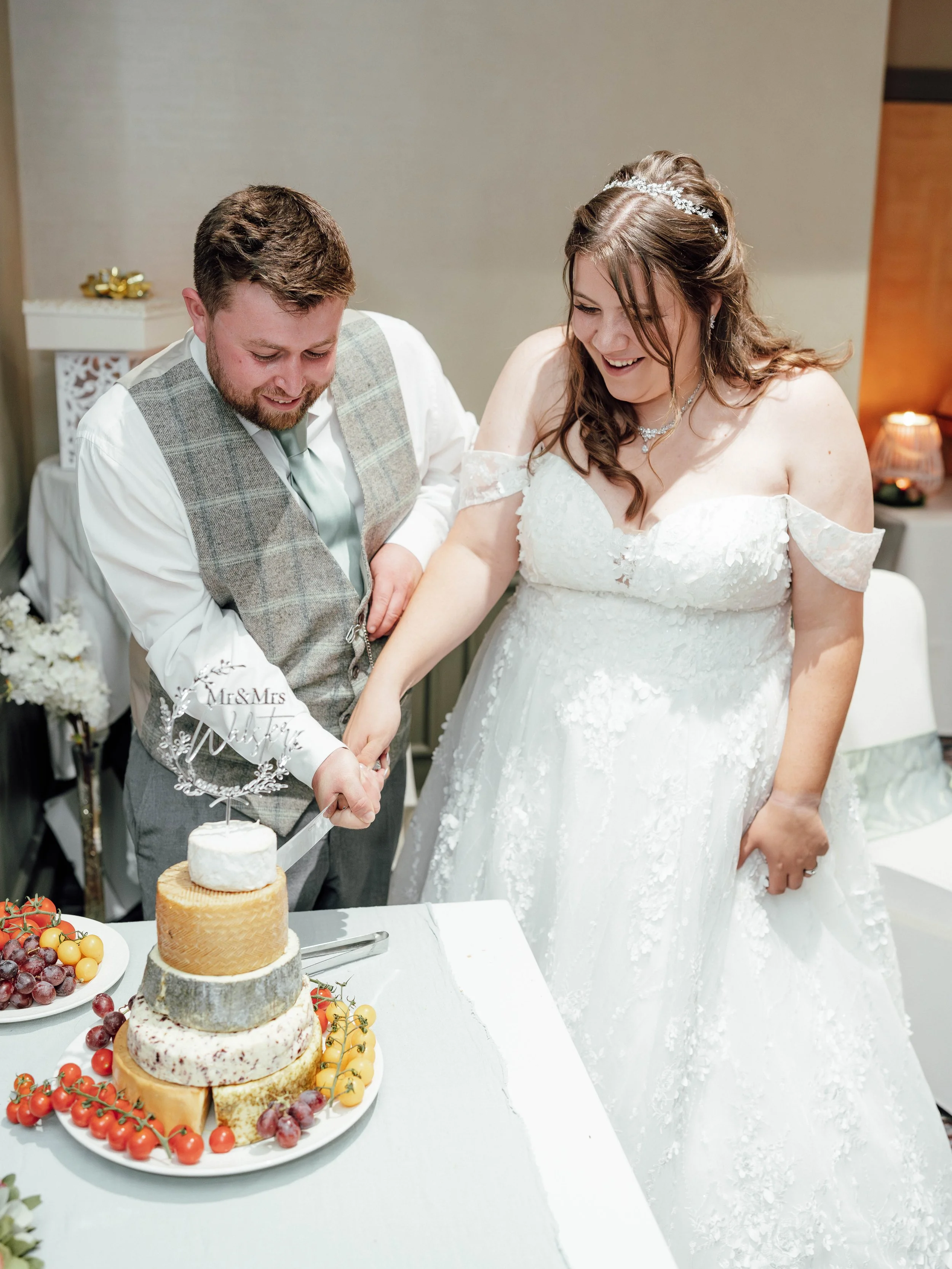 Newlyweds cutting a wedding cake, smiling, on a table with fruit and a 'Mr & Mrs' topper.