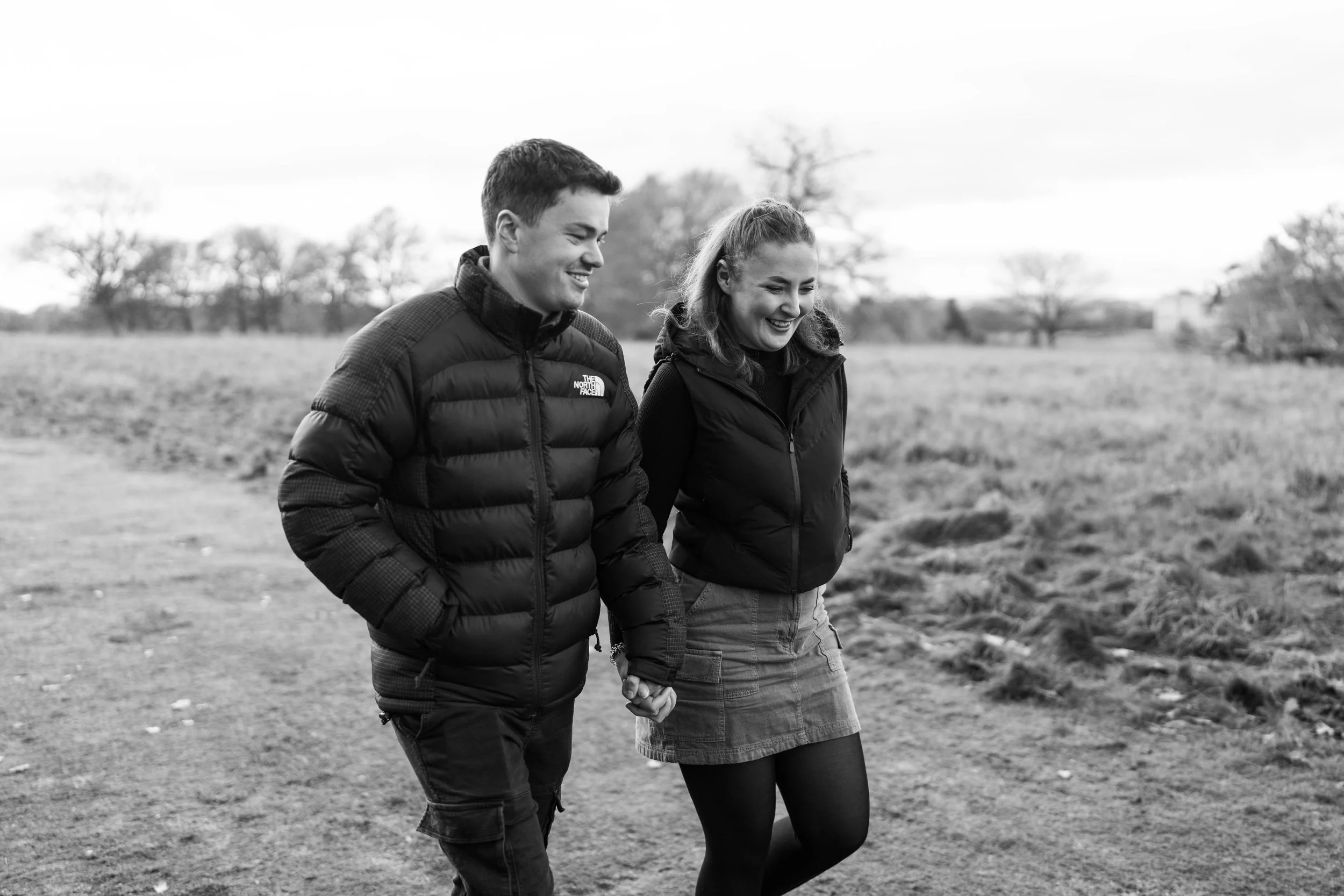 A young man and woman walking outdoors hand in hand, smiling, dressed in warm jackets, with trees and an open field in the background.