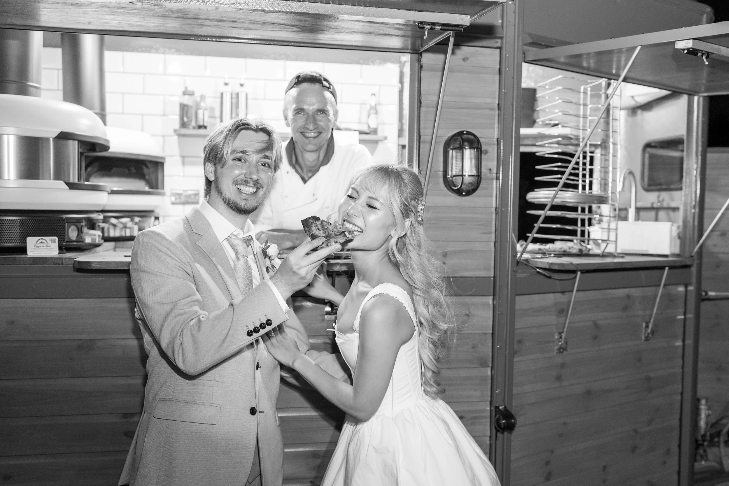A newlywed couple in wedding attire sharing pizza at a food stall with a smiling chef in the background.